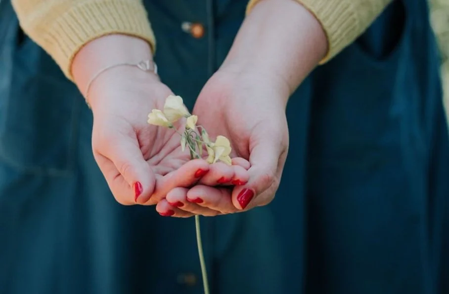 Person holding small pale yellow flowers in their hands, wearing a yellow sweater and blue dress.