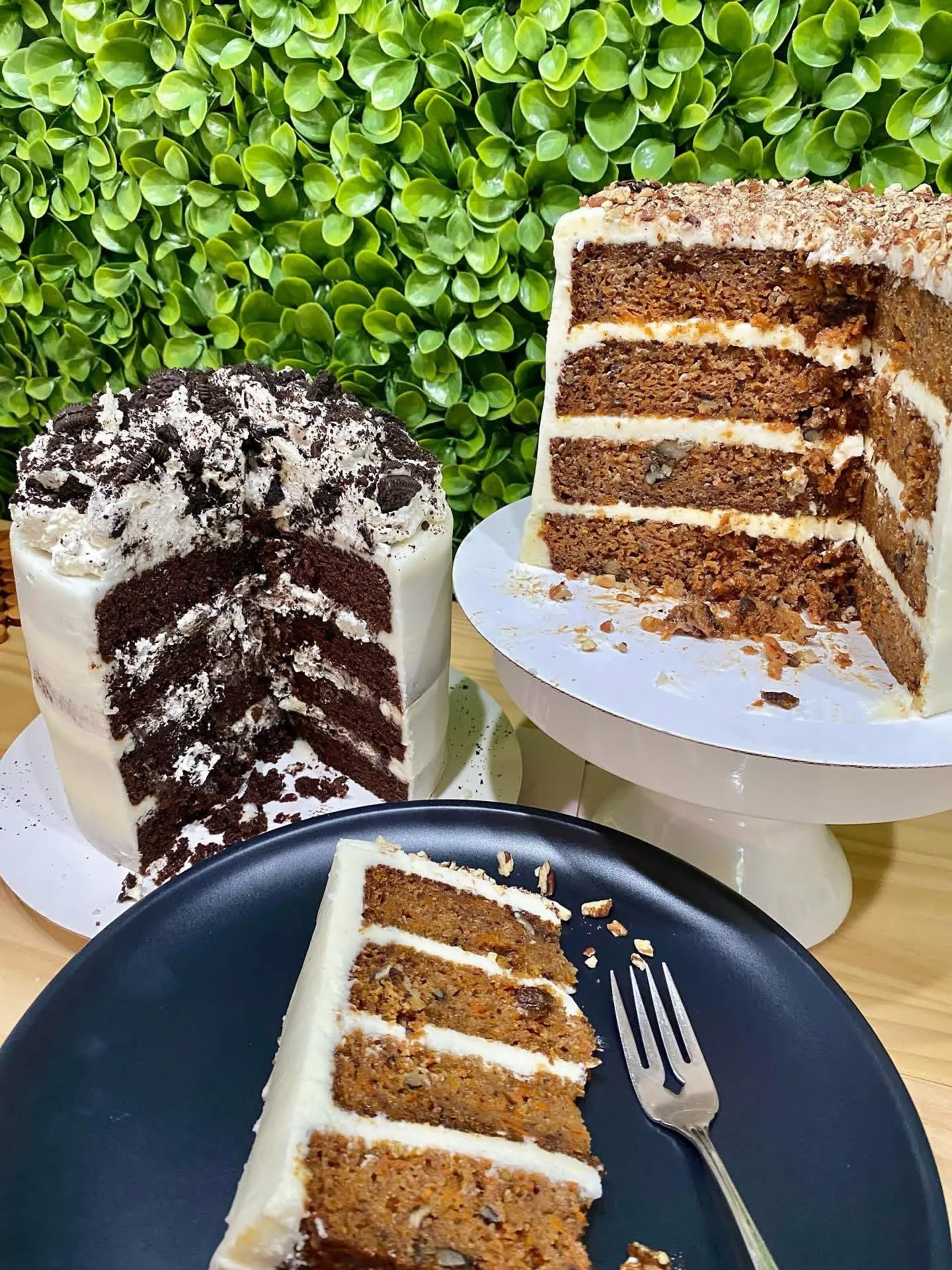 Two slices of carrot cake and one of chocolate cake on plates, with a backdrop of green leafy plants.
