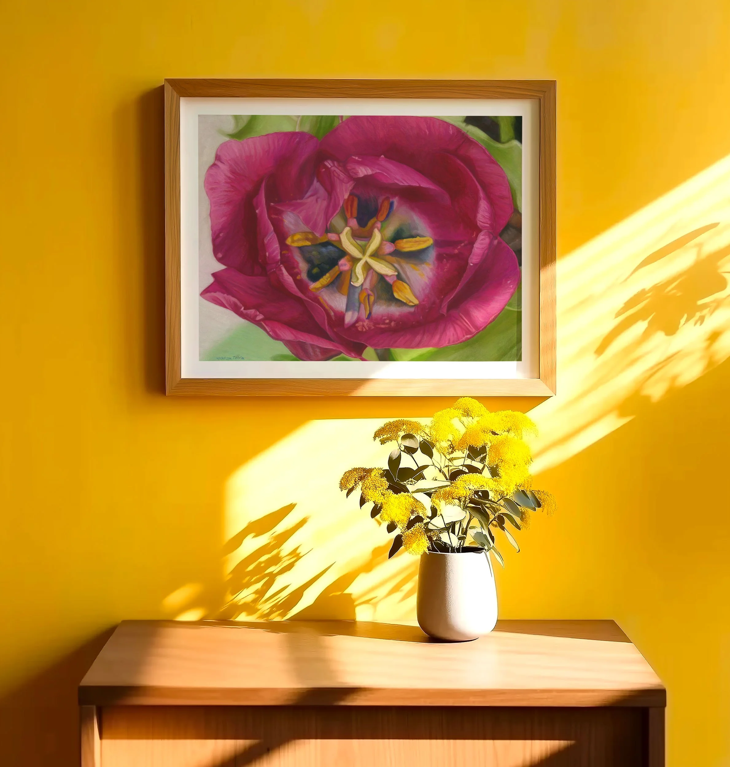 Framed magenta tulip print on a warm yellow wall above a wood table with white flowers