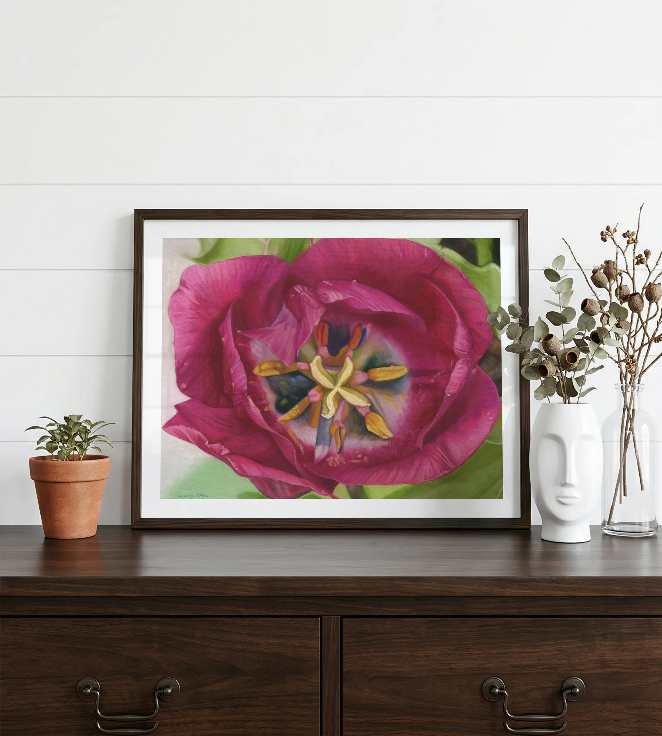 Magenta tulip art in a black frame leaning against white shiplap beside a vase of dried stems