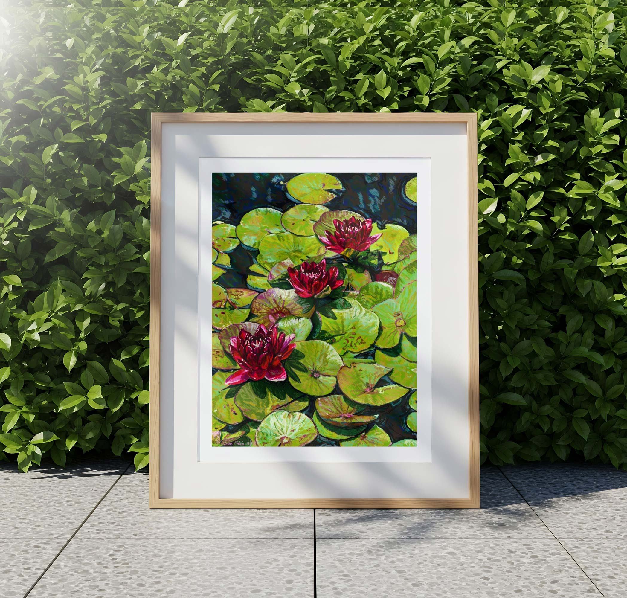 Framed red water lily print outdoors on a stone patio in front of a green hedge
