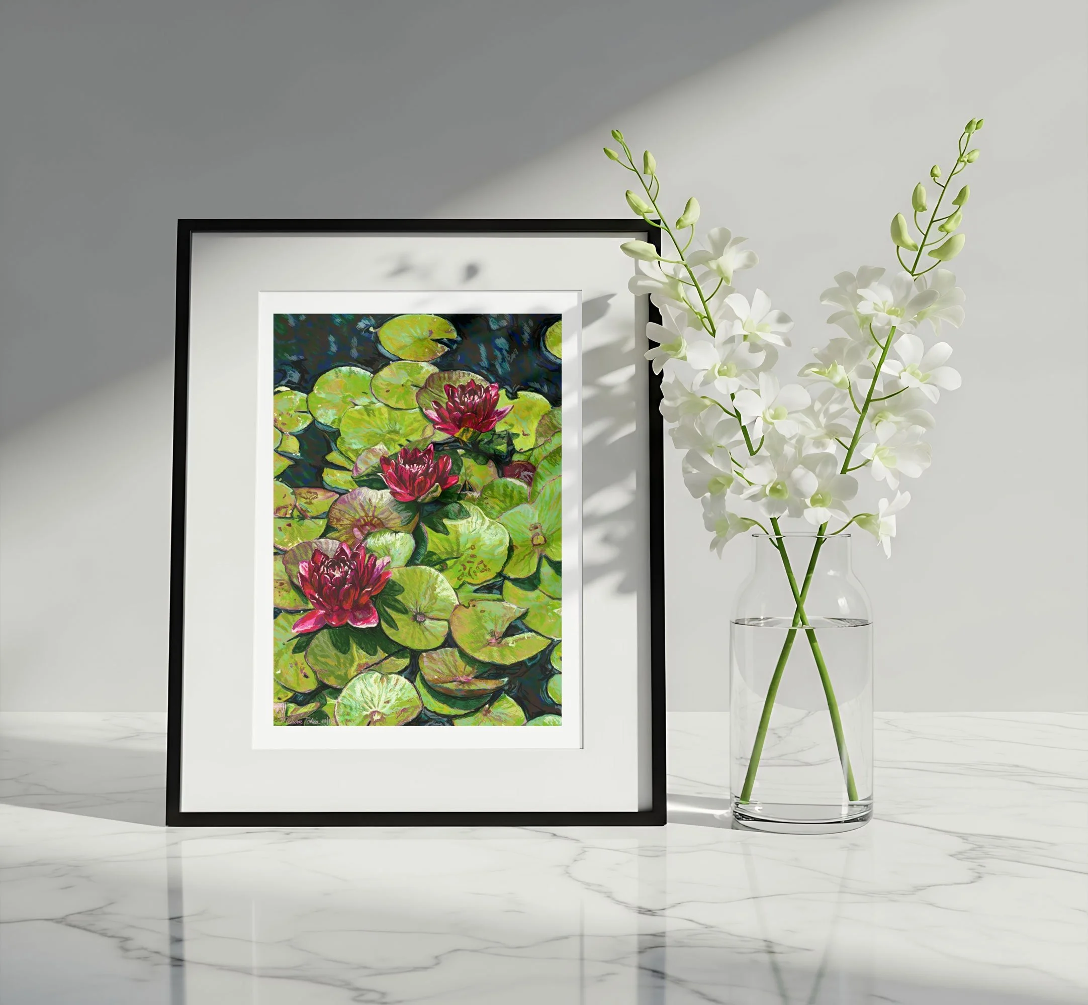 Water lily art in a black frame with a white mat beside white orchids on a marble surface