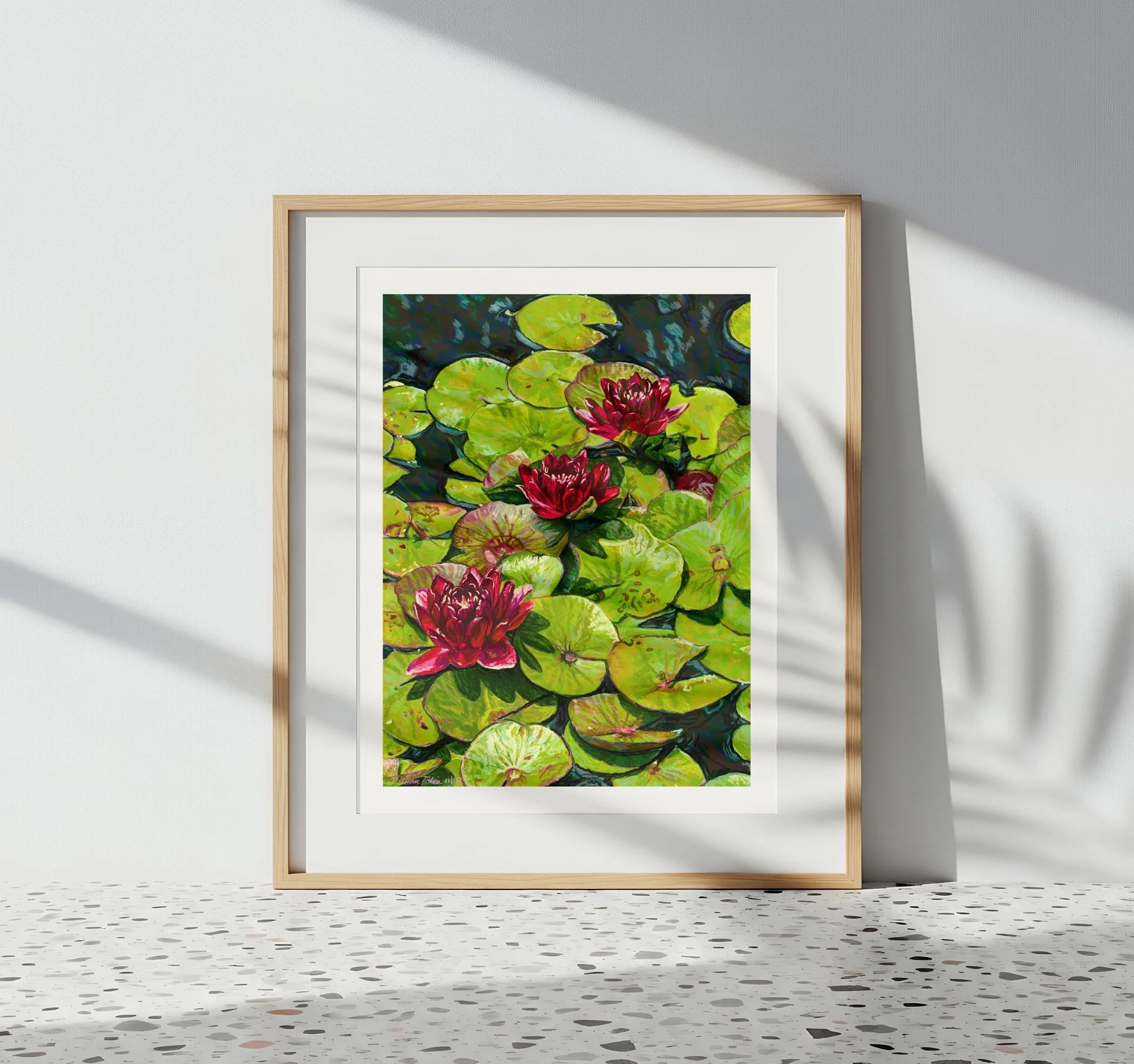Water lily art in a light wood frame leaning against a white wall with dappled leaf shadows