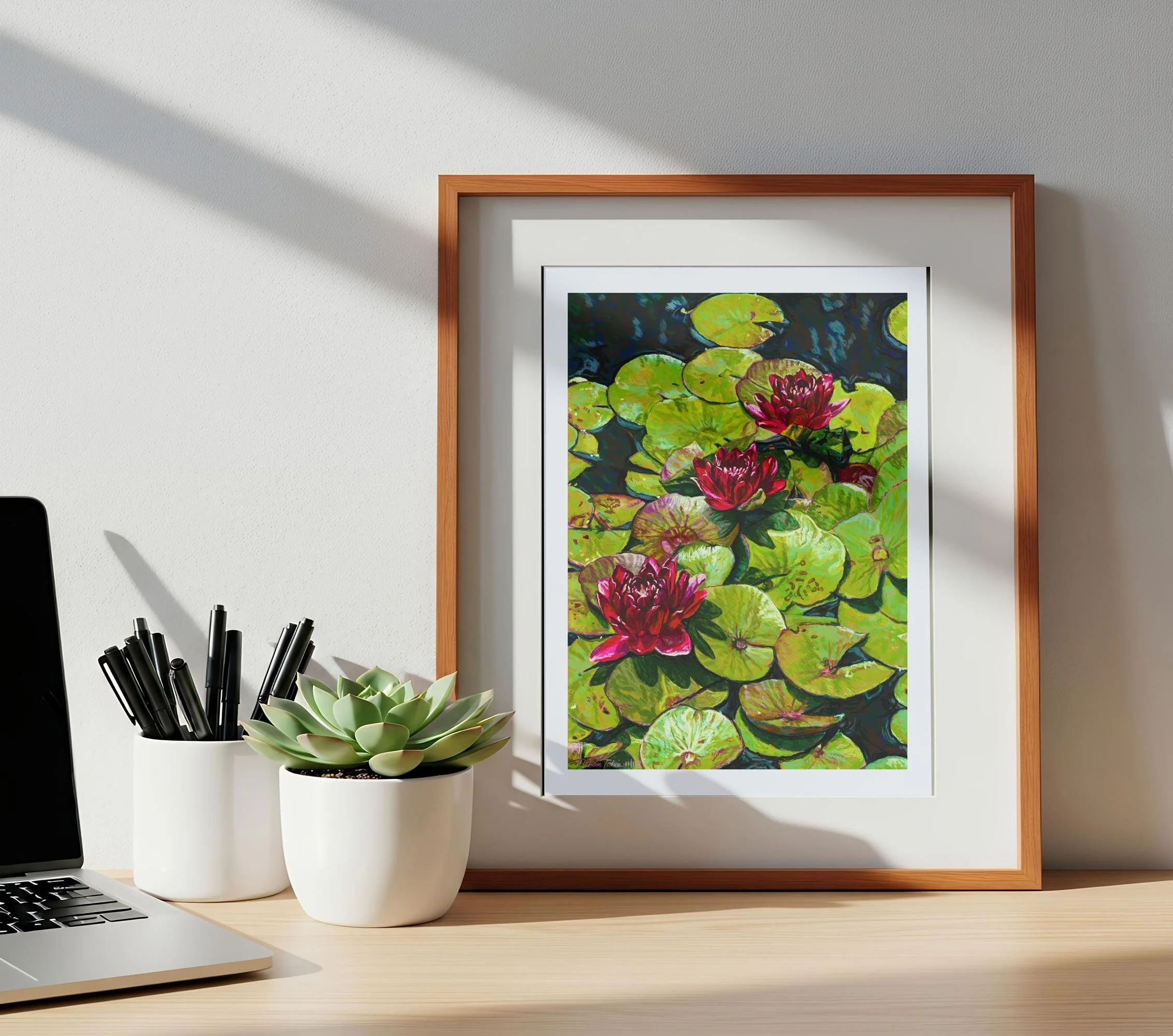 Framed lily pond print leaning on a wood desk beside a plant and pencil cup