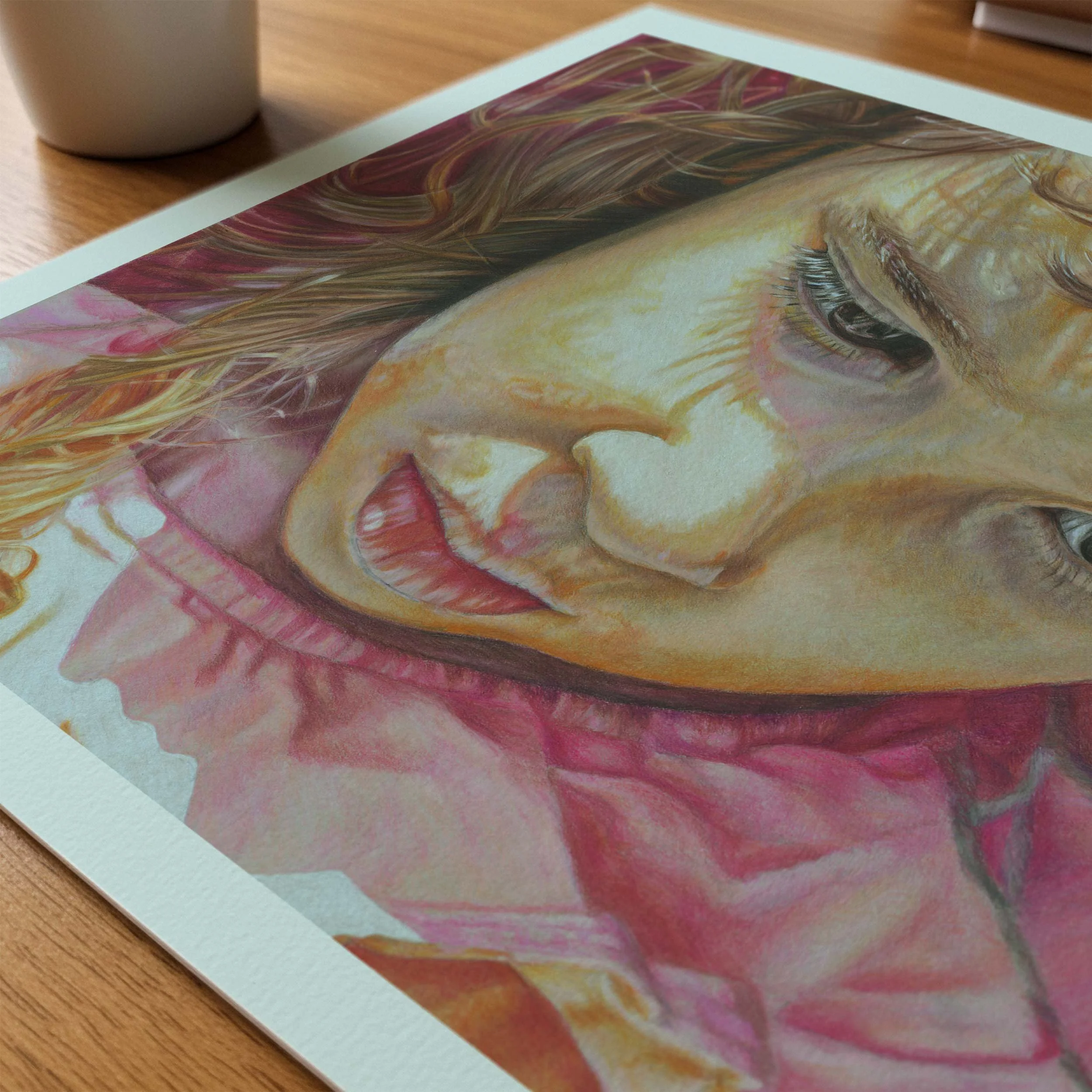 Bored colored pencil portrait print lying flat on a wood table beside a coffee mug