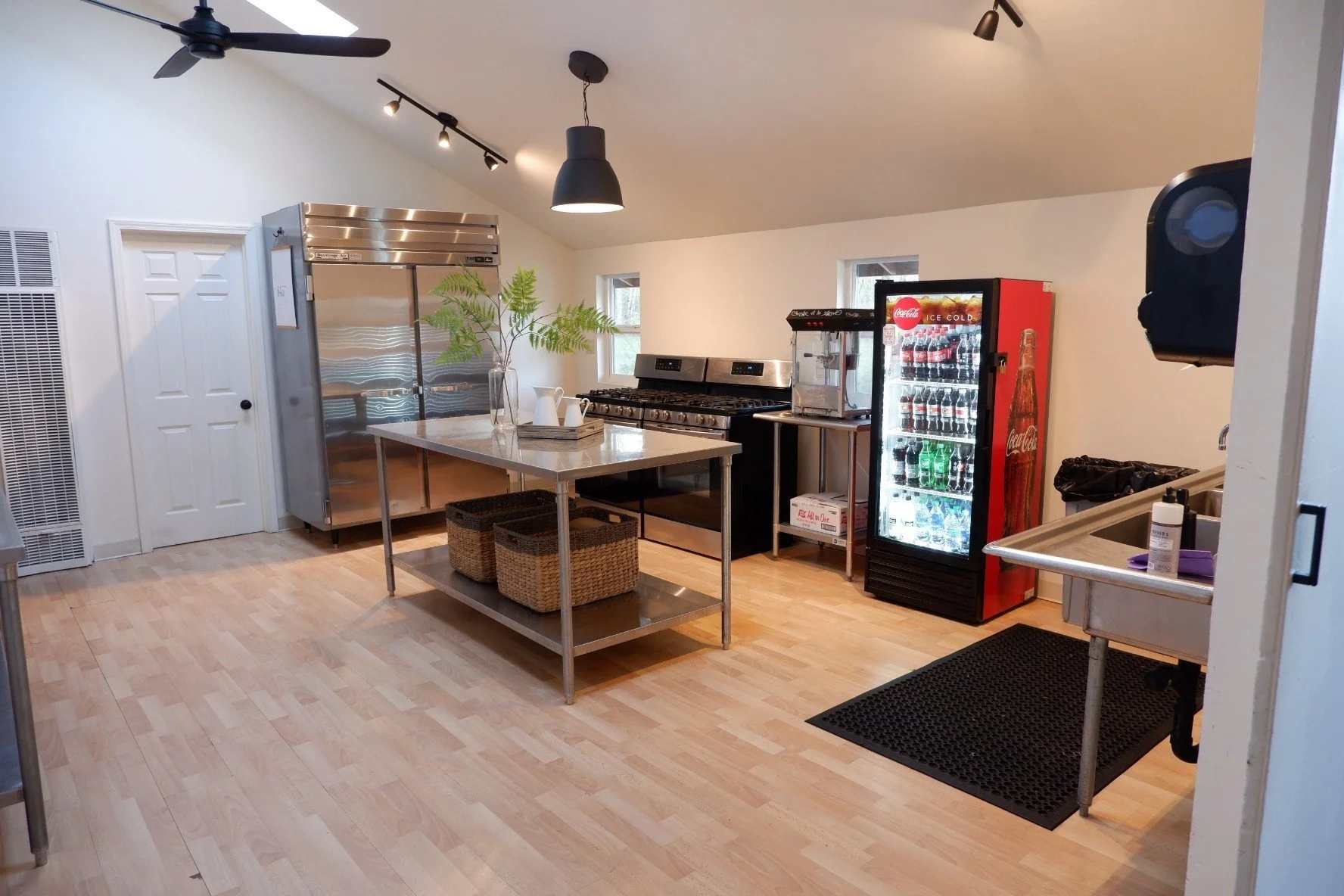 A commercial kitchen with stainless steel appliances, a white island with baskets underneath, a Coca-Cola vending machine, a soda dispenser, and a sink with cleaning supplies, illuminated by ceiling track lighting and hanging black pendant lights.