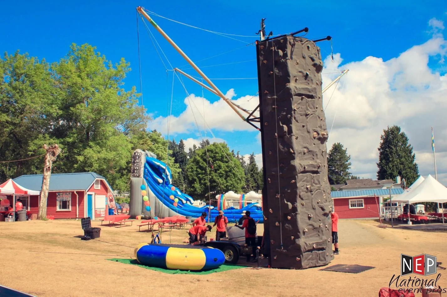 An outdoor carnival or festival with a large inflatable slide, a rock climbing wall, and various tents. Several people are gathered around, preparing or setting up the activities. There are trees and buildings in the background, and a clear blue sky with some clouds.