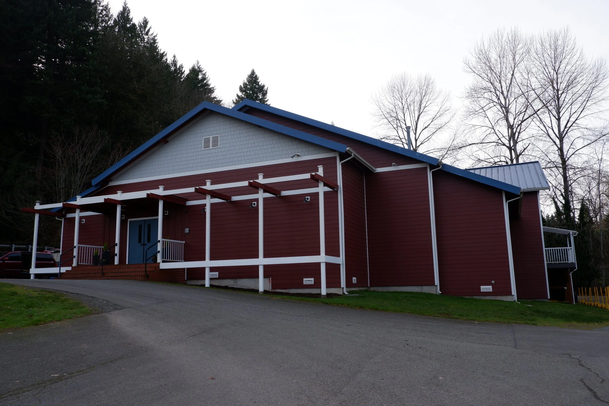 A red building with a blue roof and white trim, featuring a porch with steps and a ramp, set against a backdrop of trees and a cloudy sky.