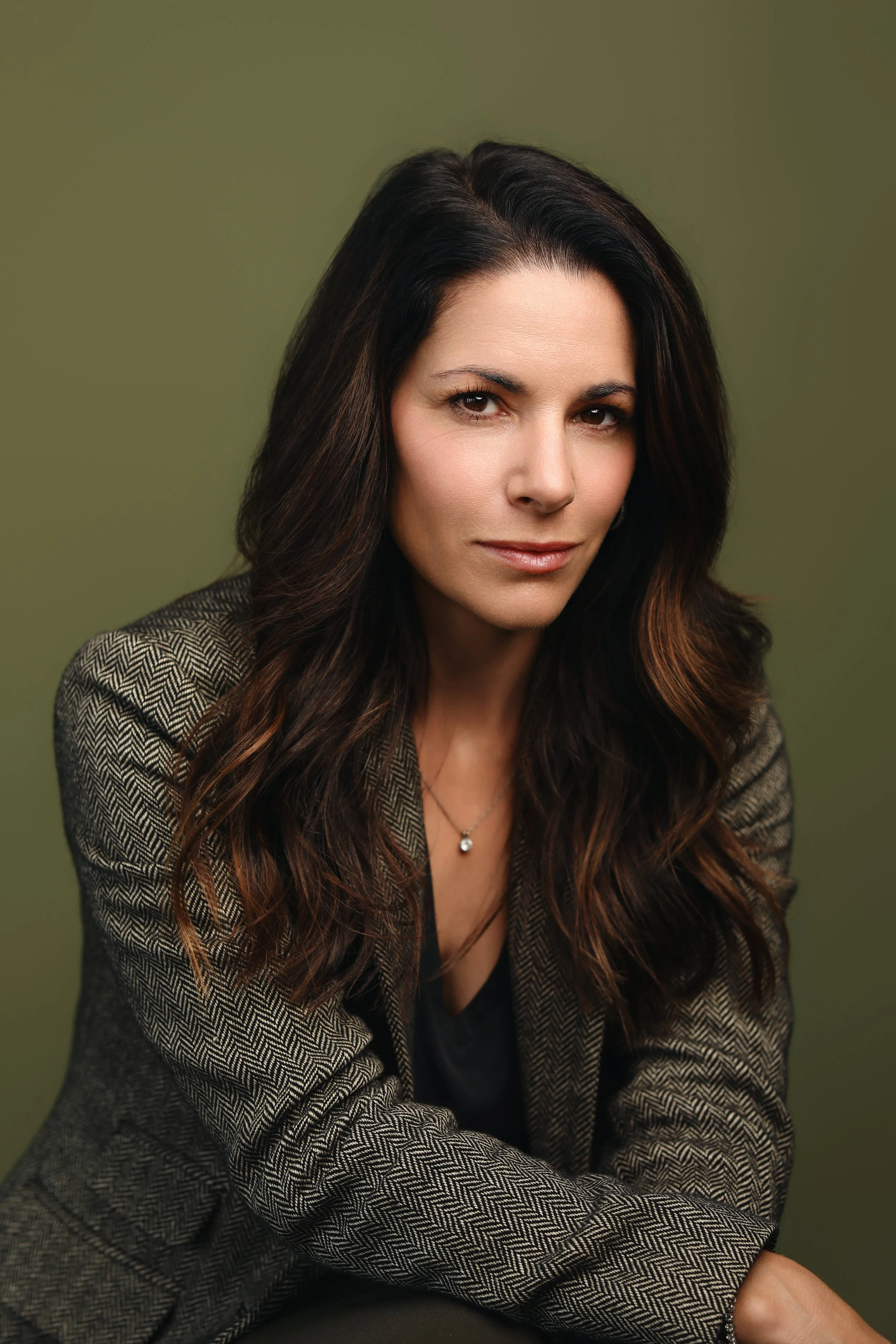 A woman with dark hair, wearing a gray blazer and jeans, sitting on a leather chair in a warmly lit room, smiling and holding a smartwatch on her wrist.