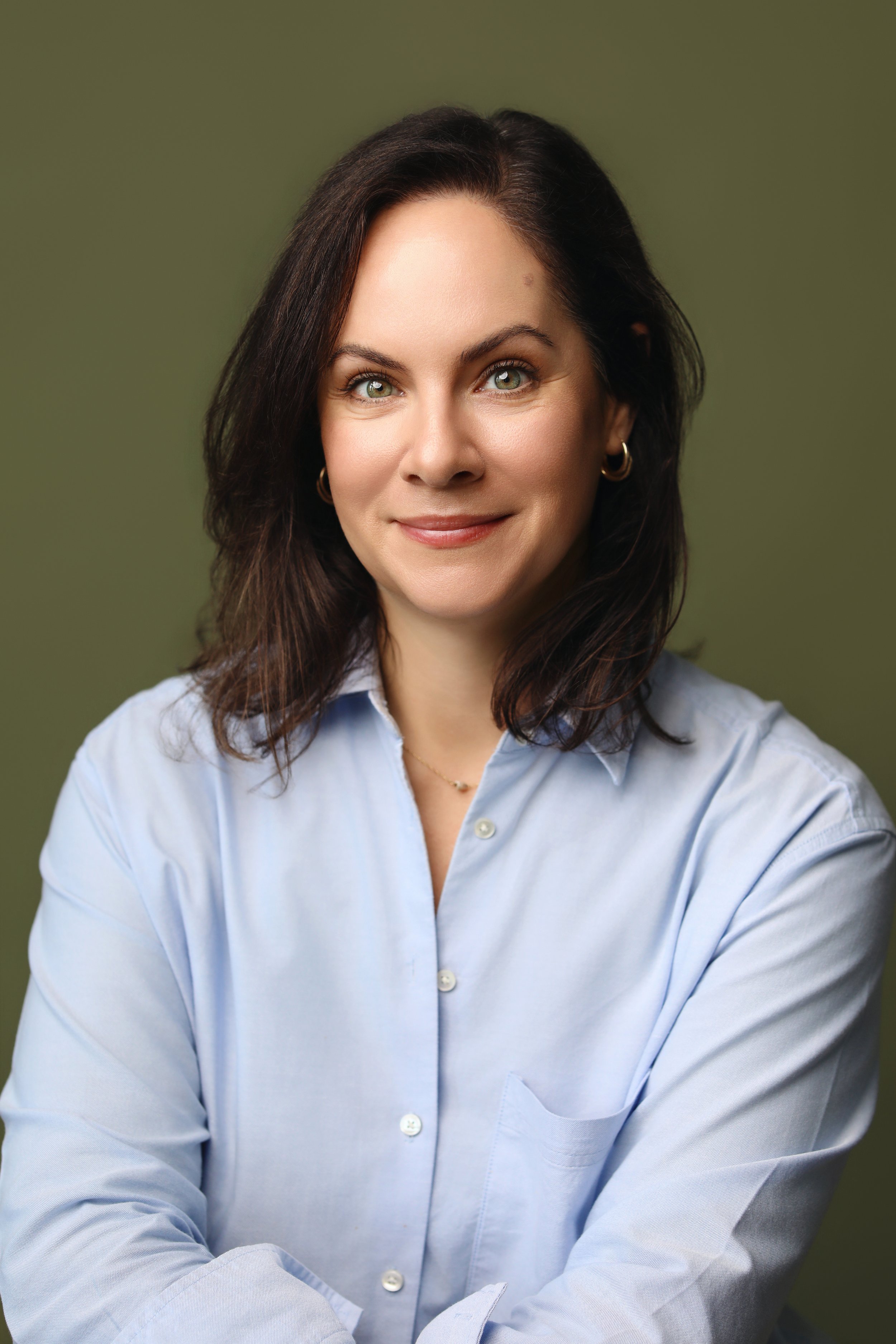 A woman with shoulder-length brown hair, wearing a white button-up shirt and blue jeans, is sitting in a brown leather chair in a cozy room with wooden wall panels and a large green leafy plant behind her. She is smiling and looking at the camera.
