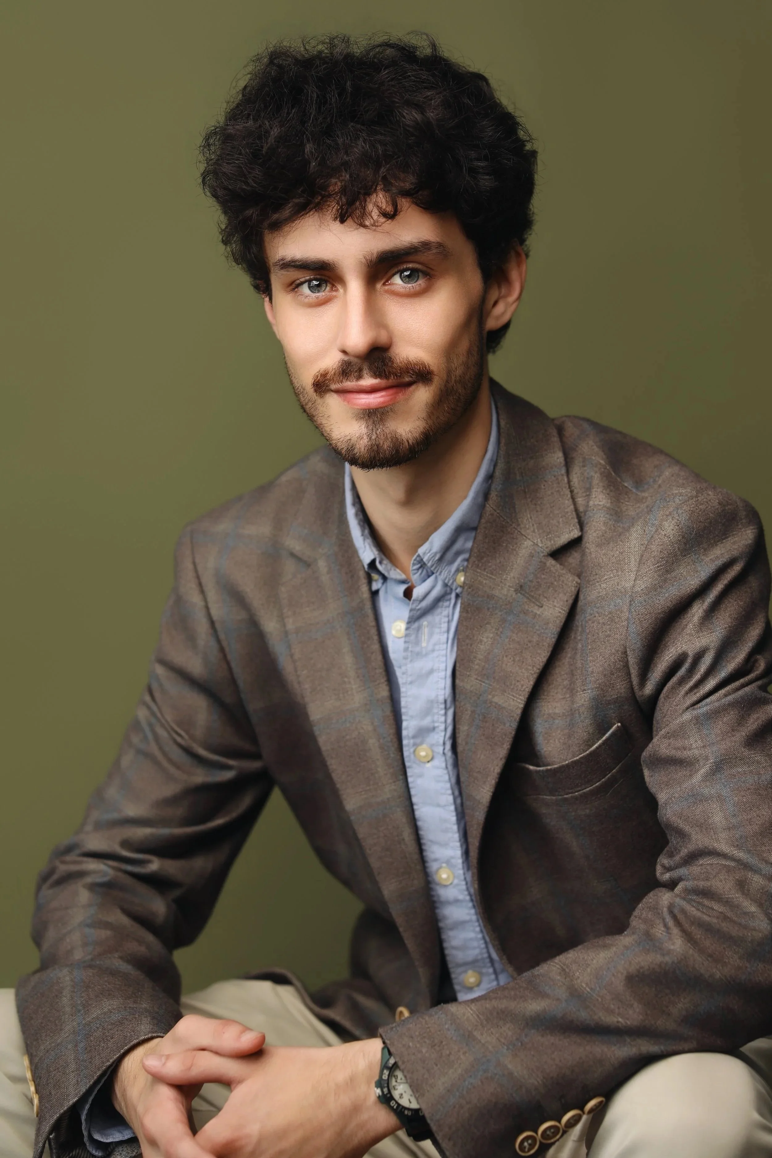 A young man with curly dark hair, a beard, and blue eyes, dressed in a brown plaid blazer, light blue shirt, and beige pants, sitting against a plain olive green background.
