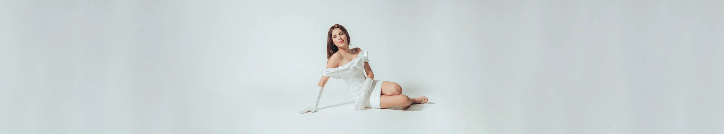 A woman with long brown hair, wearing a white off-shoulder dress and long white gloves, sitting on the floor against a plain light background.