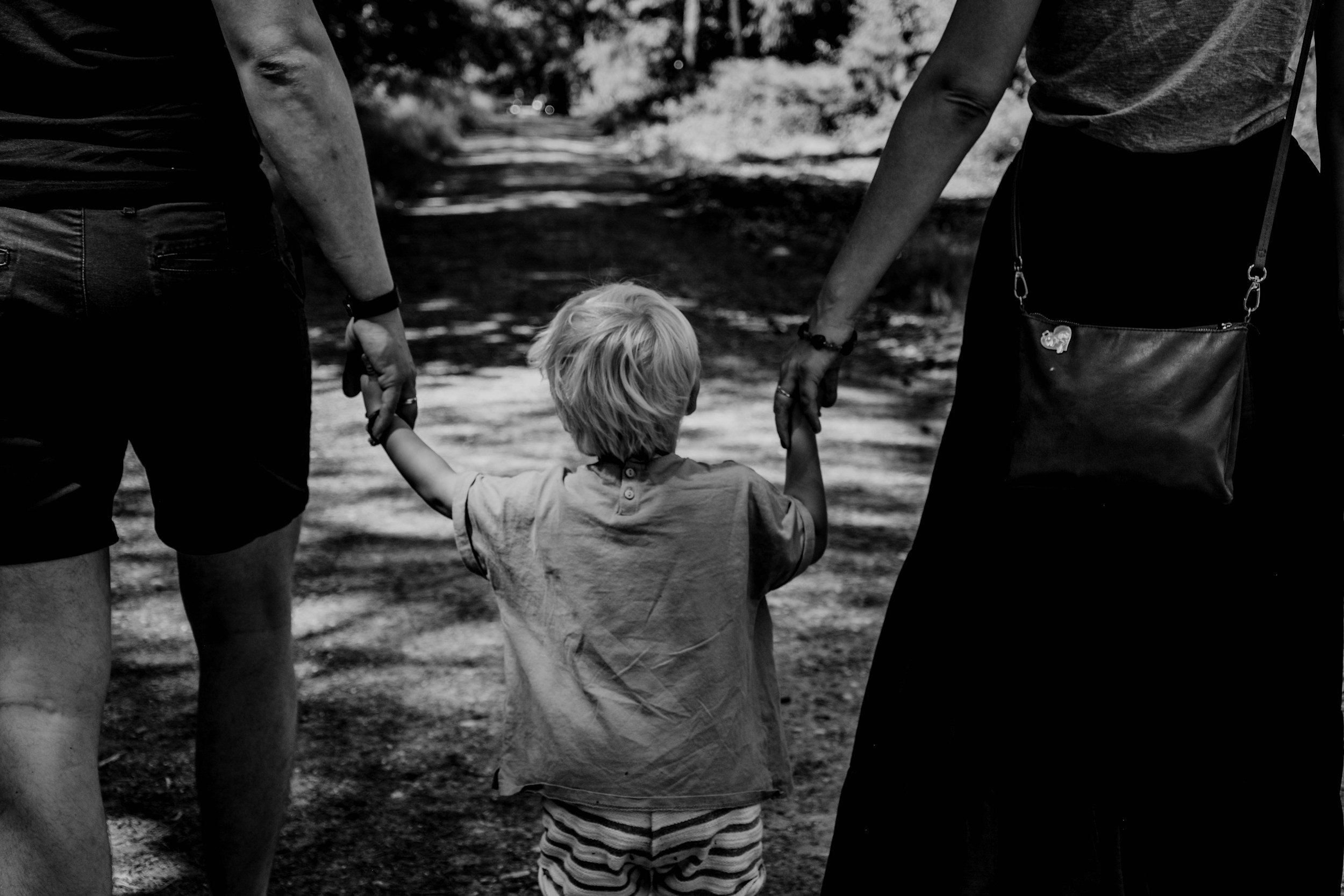 A young child holding the hands of two adults, walking on a tree-lined dirt path outdoors.