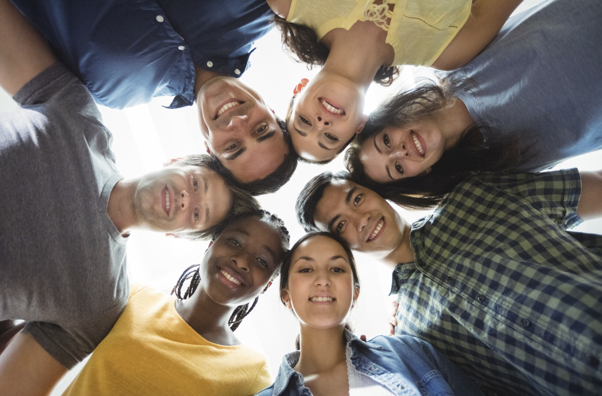 Group of diverse young adults standing in a circle and looking down at the camera, smiling.