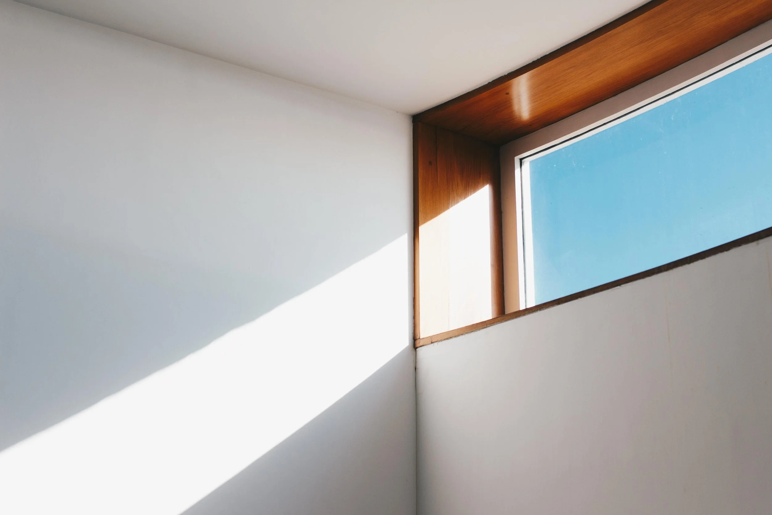 Interior corner of a room with a large rectangular window, wooden trim, and bright sunlight creating a shadow on the white wall.