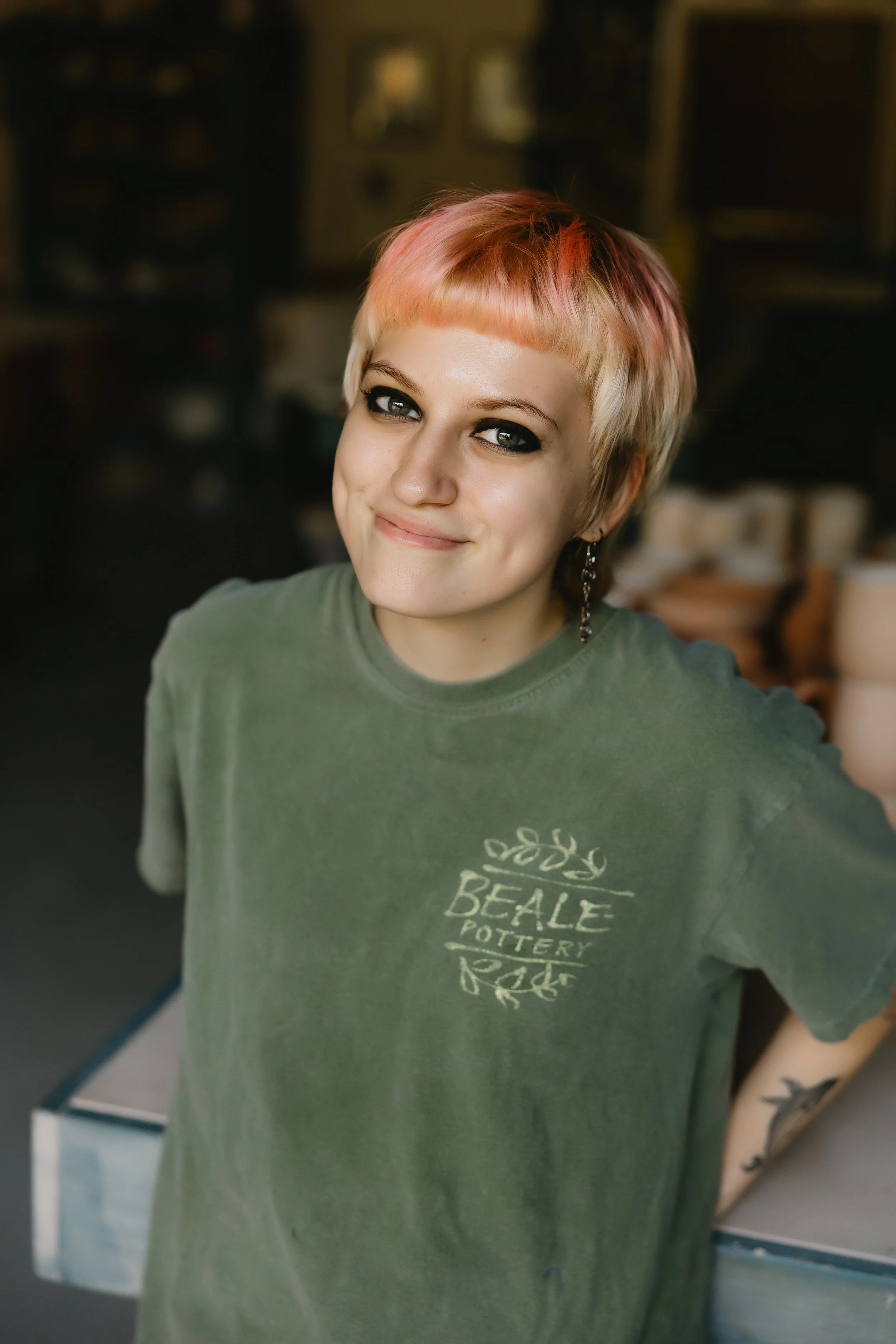 A young woman with short, light pink hair smiling at the camera in a pottery studio. She's wearing a green Beale Pottery t-shirt and has a tattoo on her right forearm.