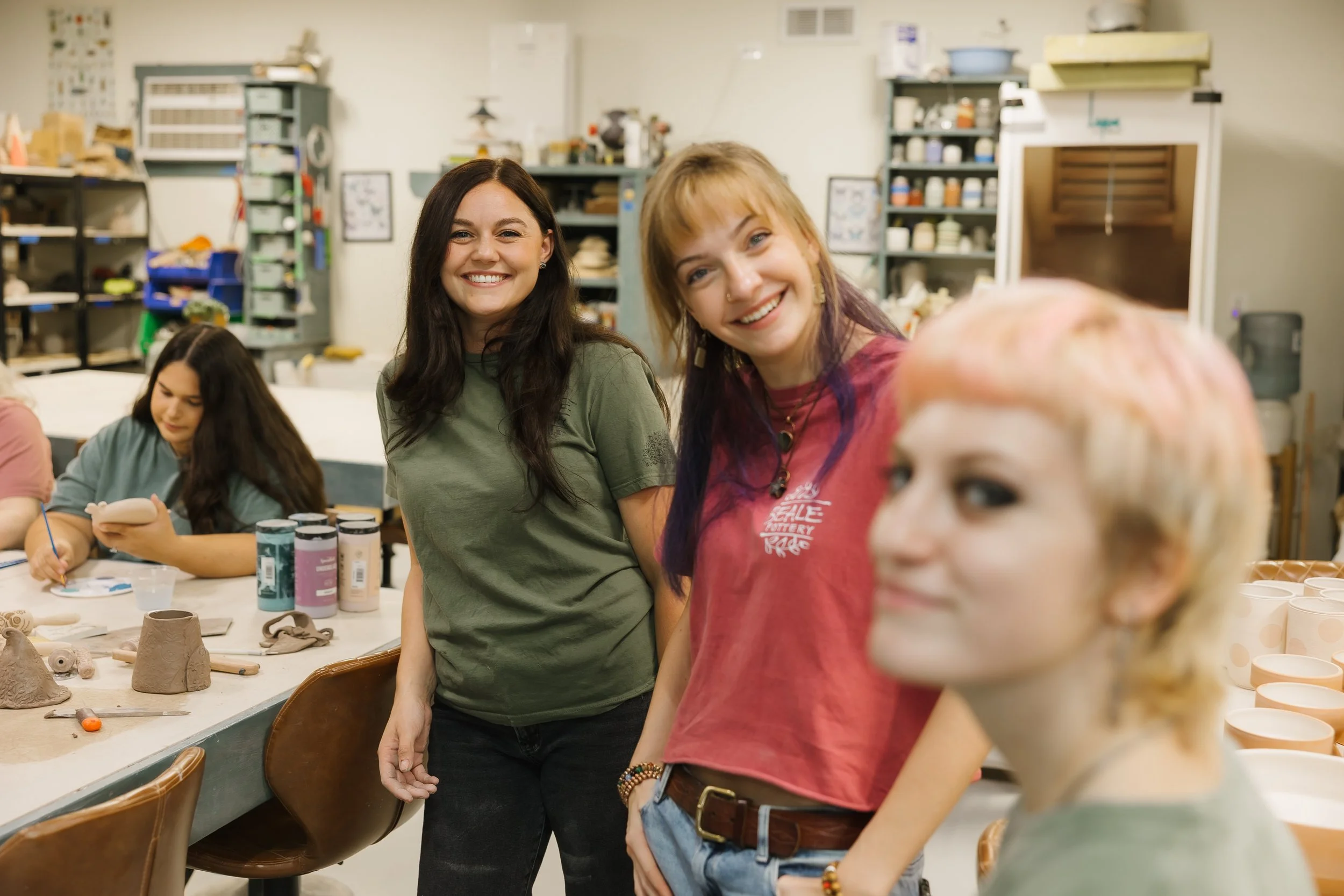 Three women smiling in a pottery studio with shelves of supplies and pottery on the table.
