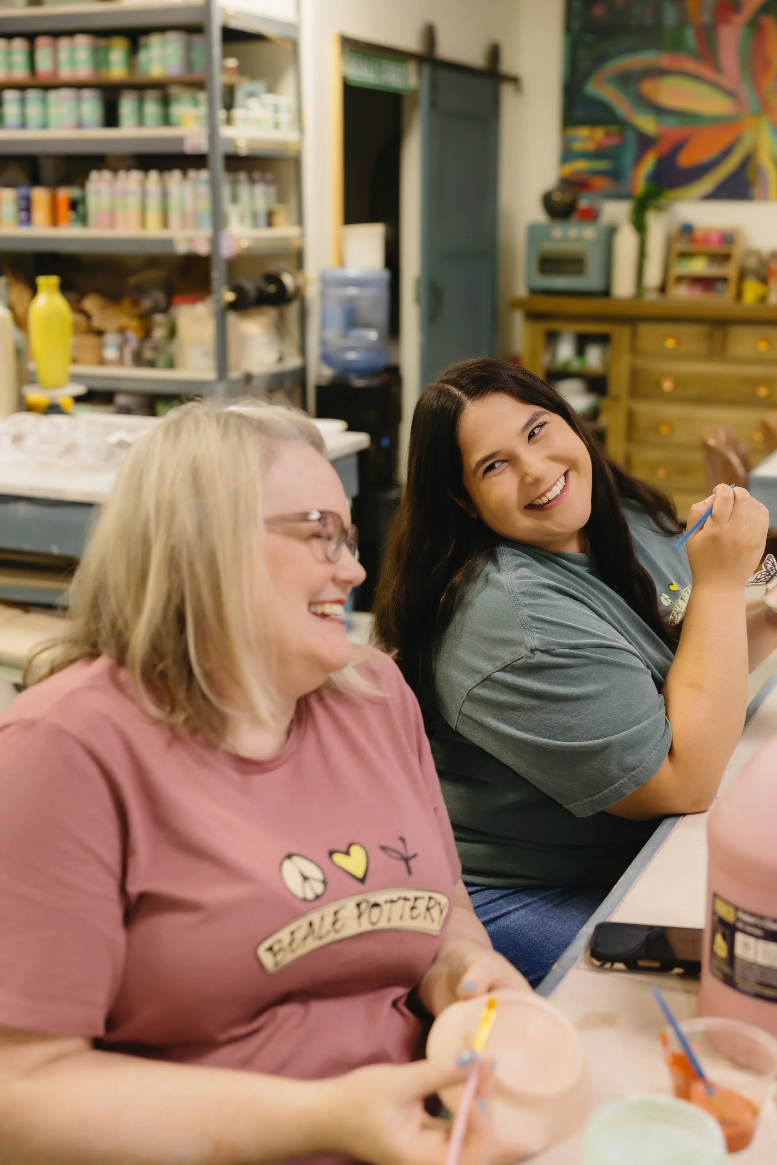 Two women sitting at a table, smiling and enjoying a pottery activity in a pottery studio with shelves of supplies in the background.