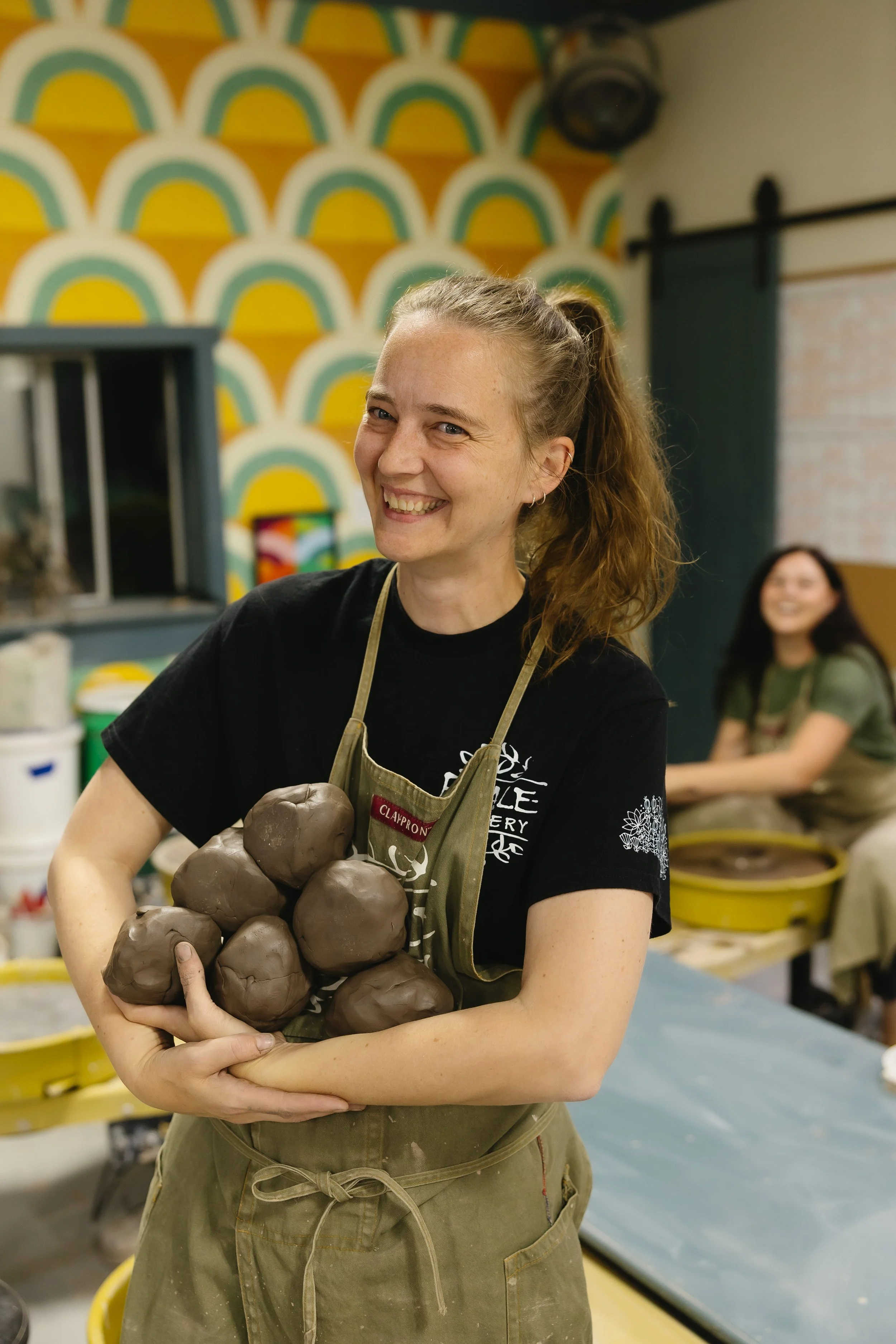 A smiling woman in a black t-shirt and green apron holding clay sculptures in a pottery class.
