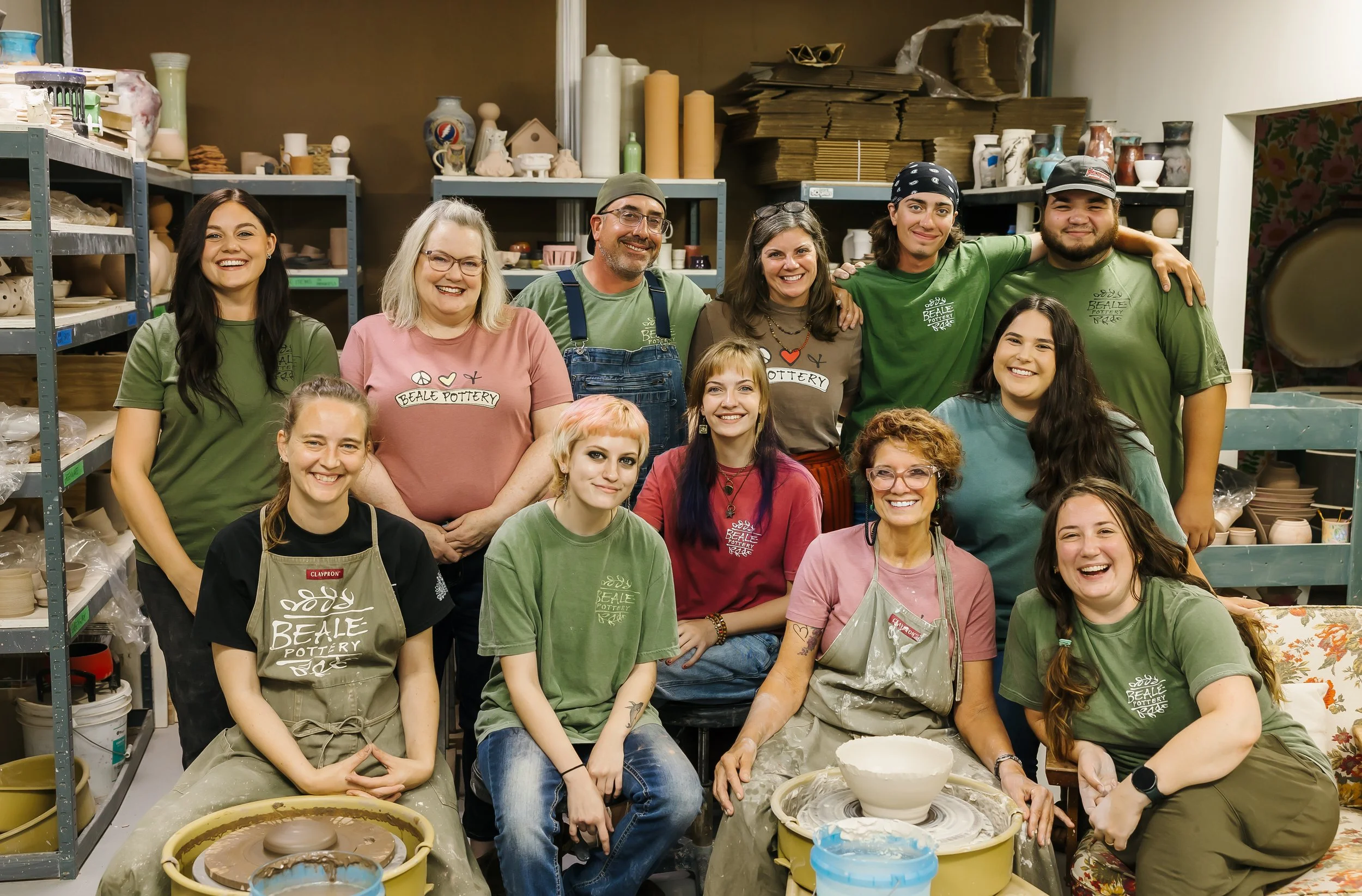 Group of people in a pottery studio, smiling and posing for the photo, with shelves of pottery supplies and finished pottery in the background.