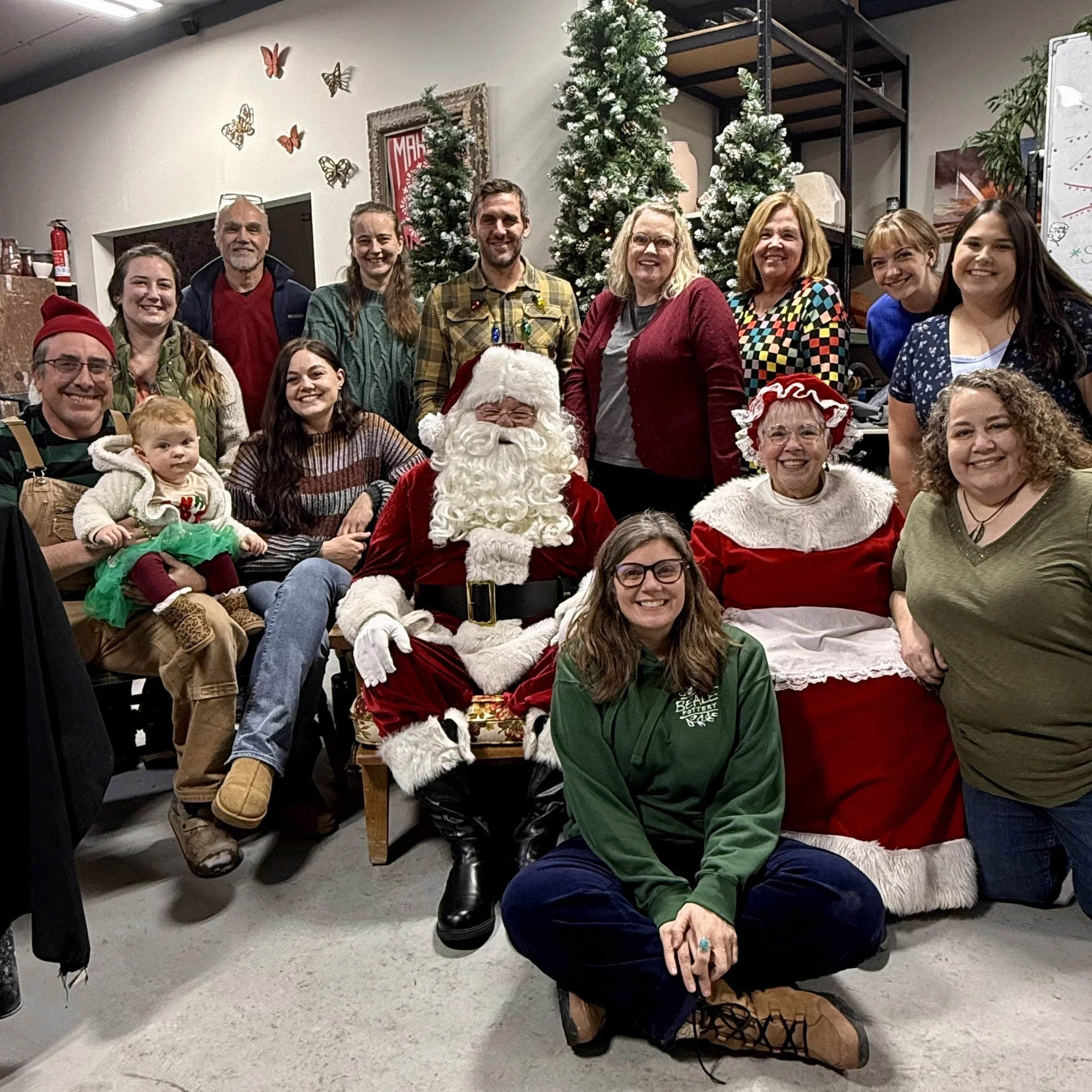 Group of people gathered around Santa Claus and Mrs. Claus at a Christmas party, with decorated trees and holiday decor in the background.