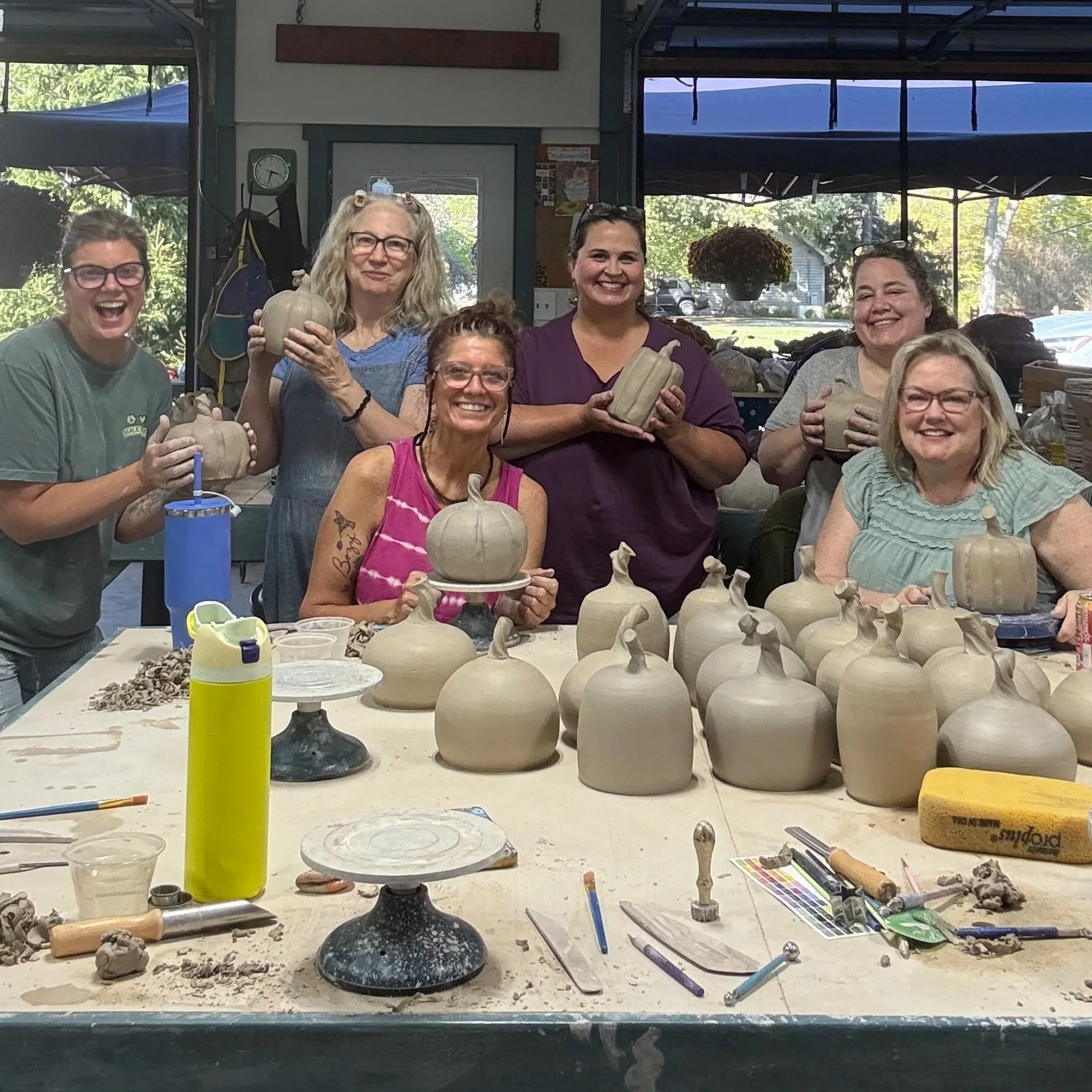 Group of six women in a pottery studio holding finished ceramic pumpkins, surrounded by unglazed pumpkin-shaped ceramics on the table, with pottery tools and supplies visible.