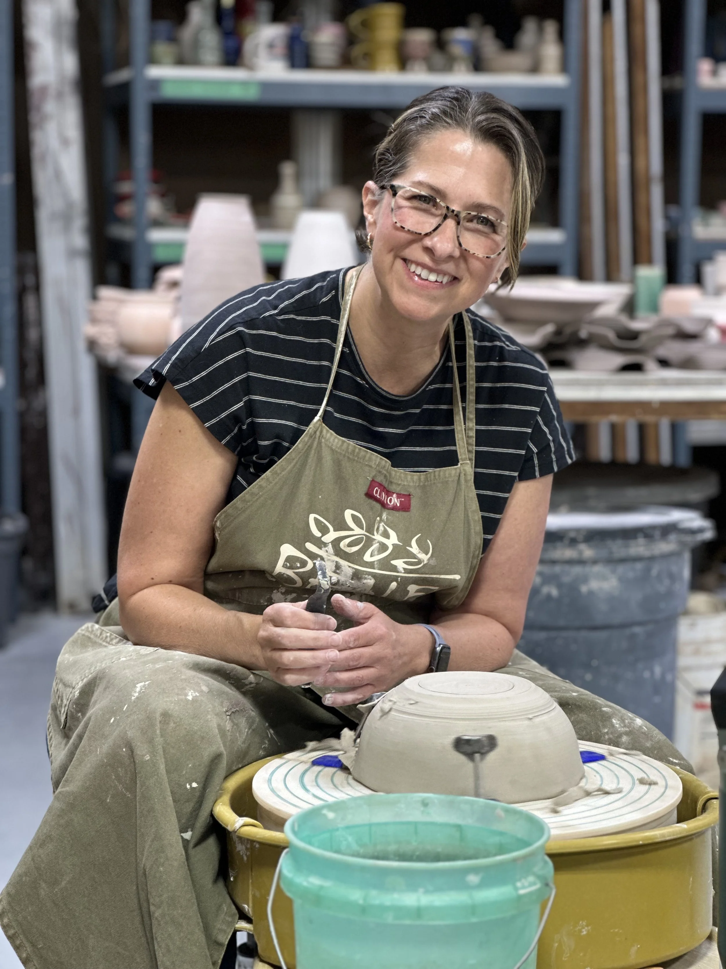 Woman smiling at pottery wheel in pottery studio, working on a ceramic piece, wearing glasses and apron.