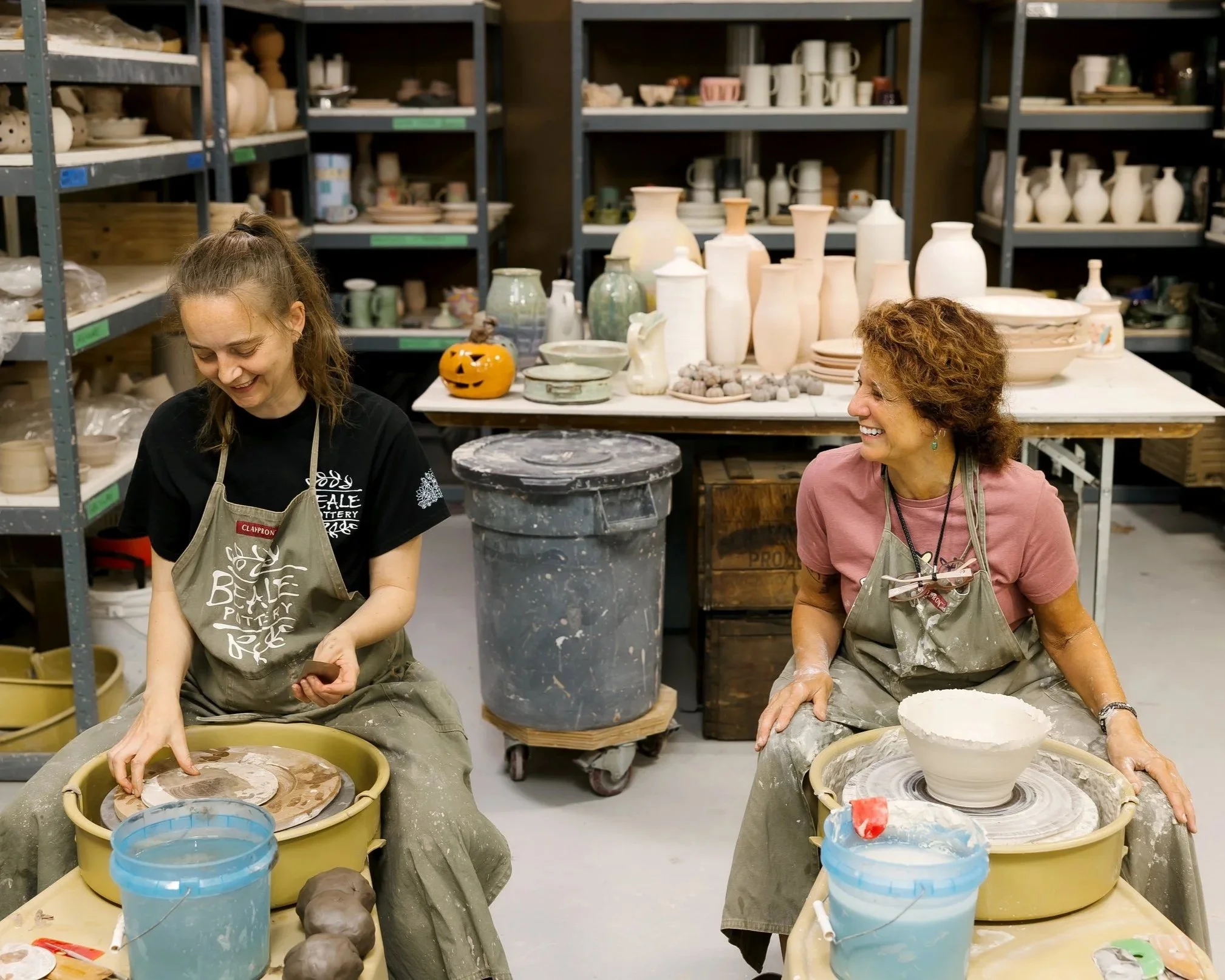Two women working together on pottery projects in a ceramics studio. The woman on the left, with long hair, is focused on shaping clay on a wheel, while the woman on the right, with curly hair, is smiling and working on her own pottery project. Shelves filled with pottery and ceramics are in the background.