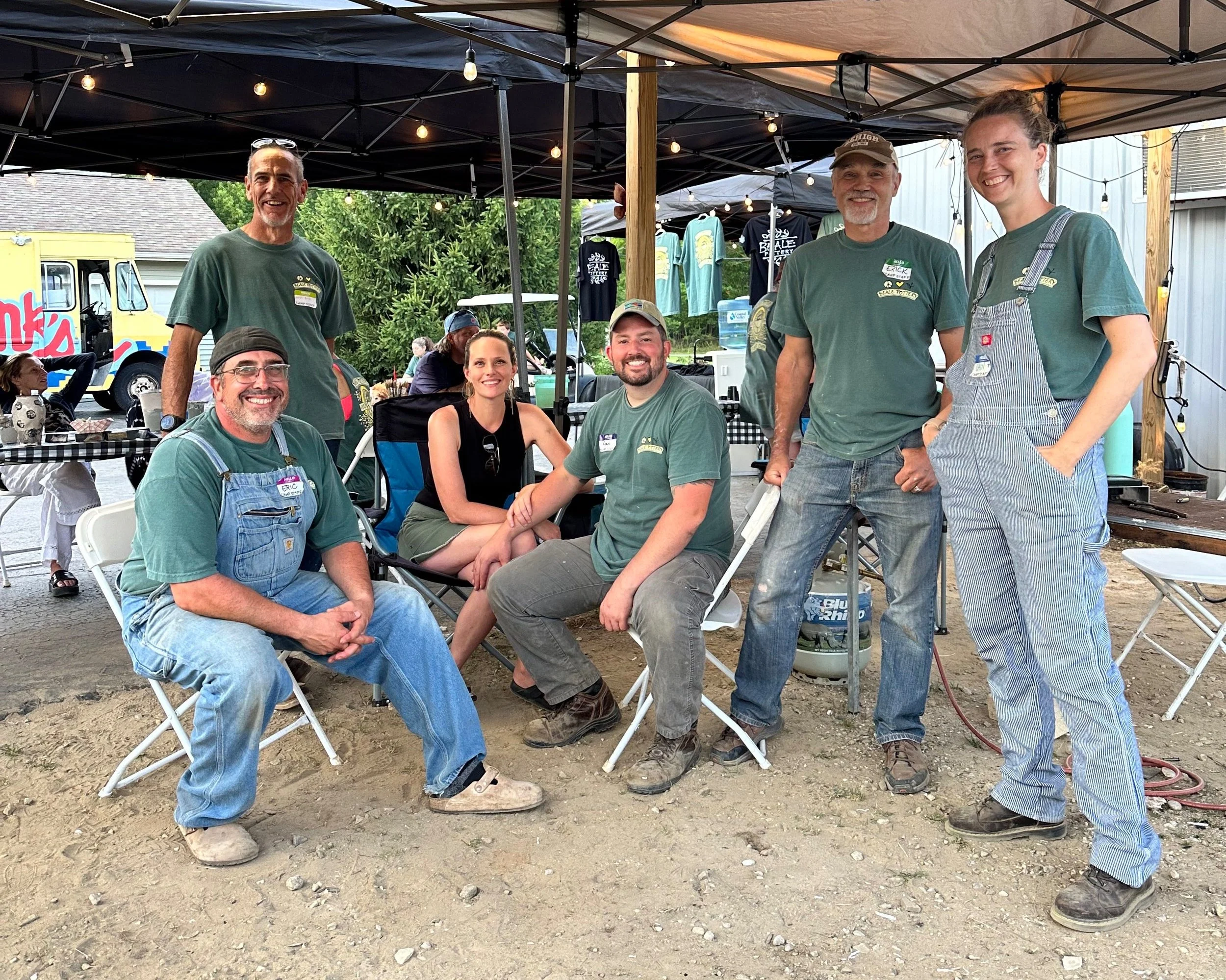 Group of seven people, some sitting and some standing, at an outdoor event under a canopy. They are smiling and appear relaxed, with some wearing green shirts and overalls, and others in casual clothing. There are tents, tables, and a food truck in the background.