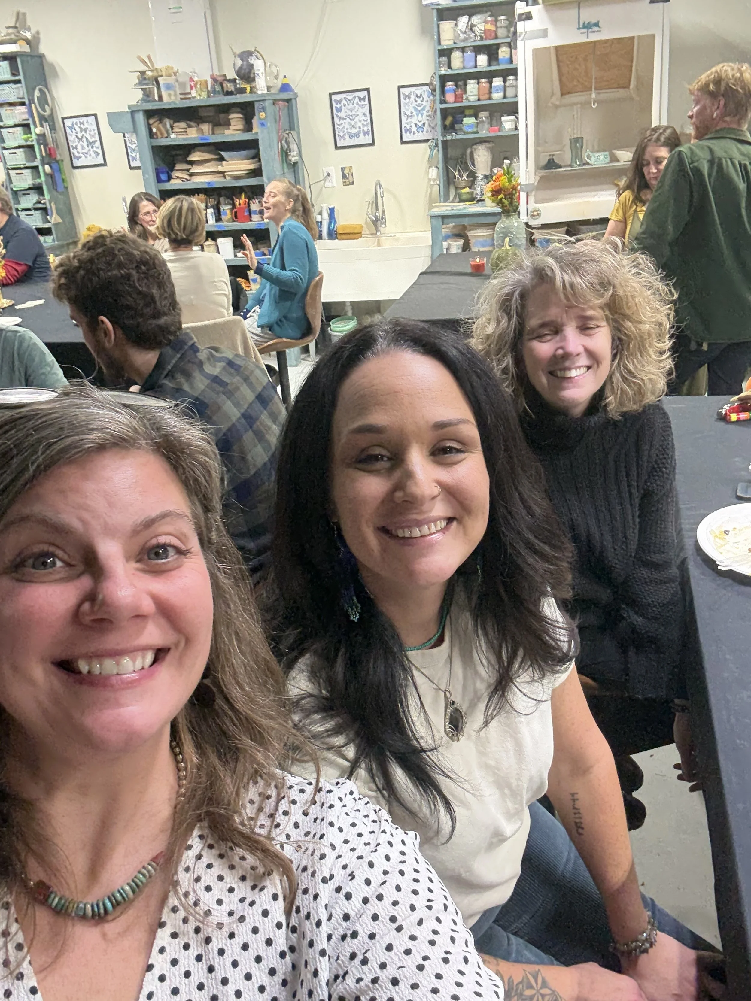 Group of people smiling and taking a selfie at a gathering in a cozy, decorated kitchen or art studio with shelves, artwork, and art supplies in the background.