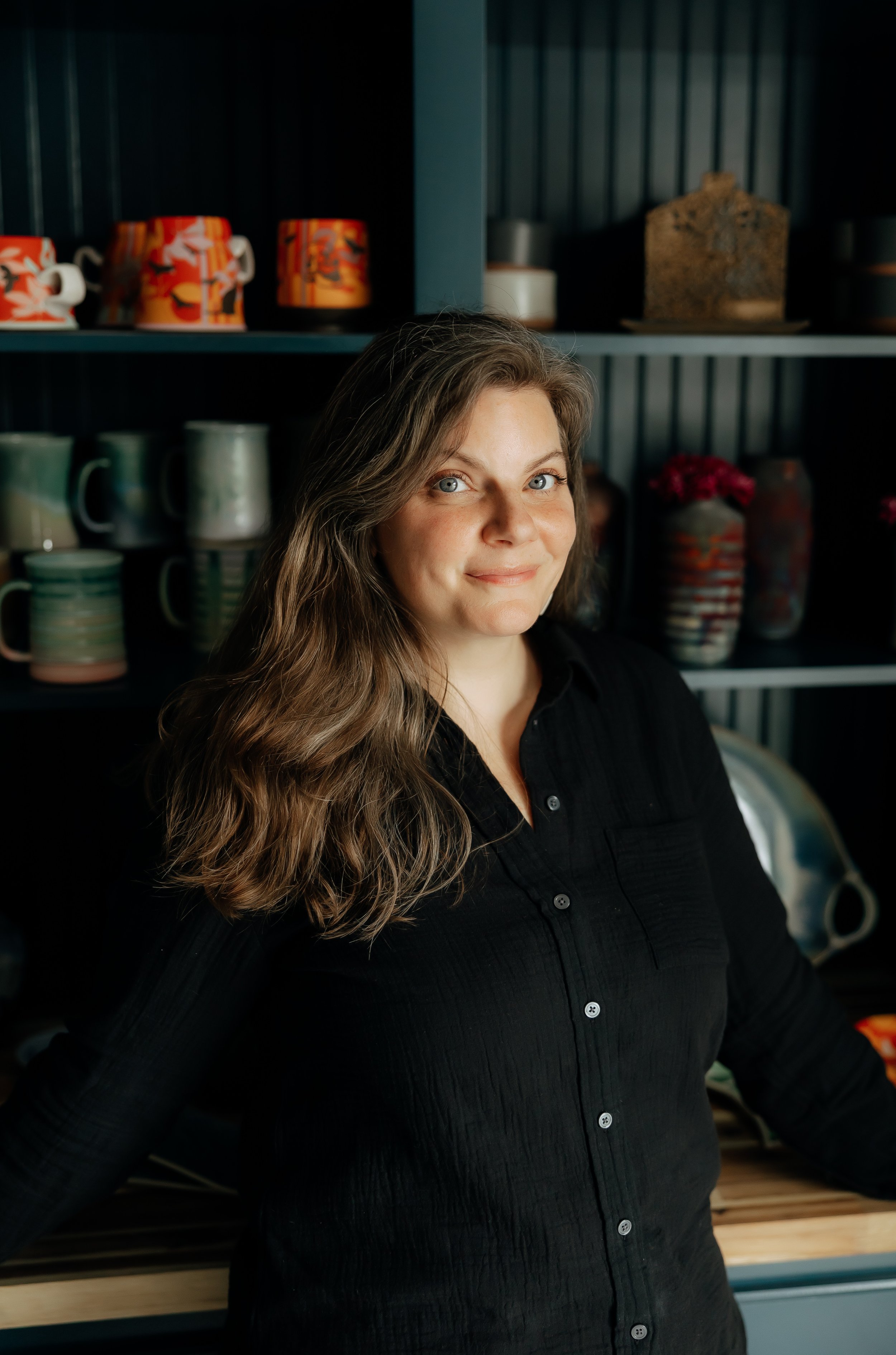 A woman with long wavy brown hair and blue eyes smiling while standing in front of shelves with colorful cups, mugs, and vases in a kitchen or store setting.
