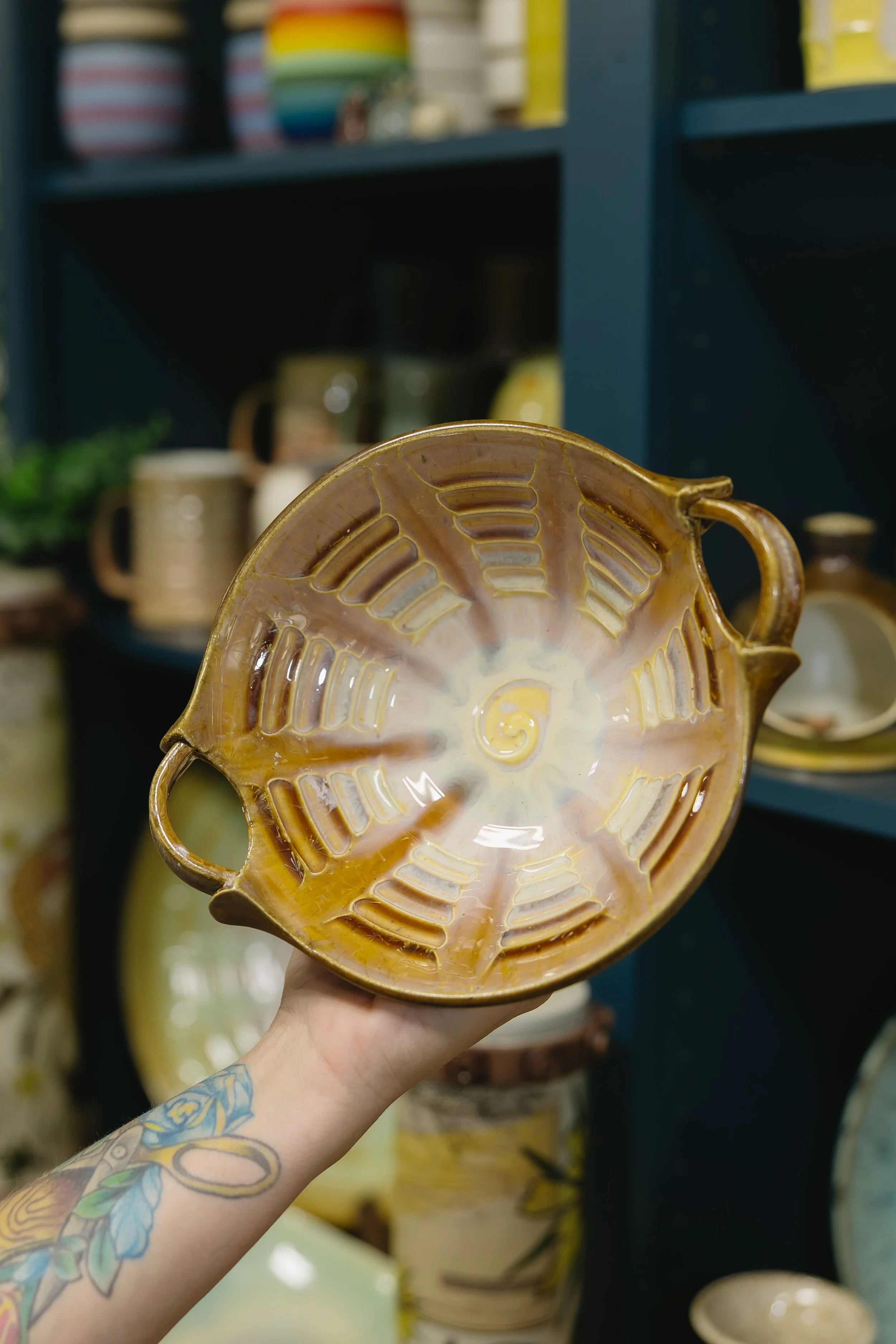 Person holding a yellow ceramic vessel with open side handles, inside a pottery or gift shop with shelves of mugs, bowls, and other pottery.