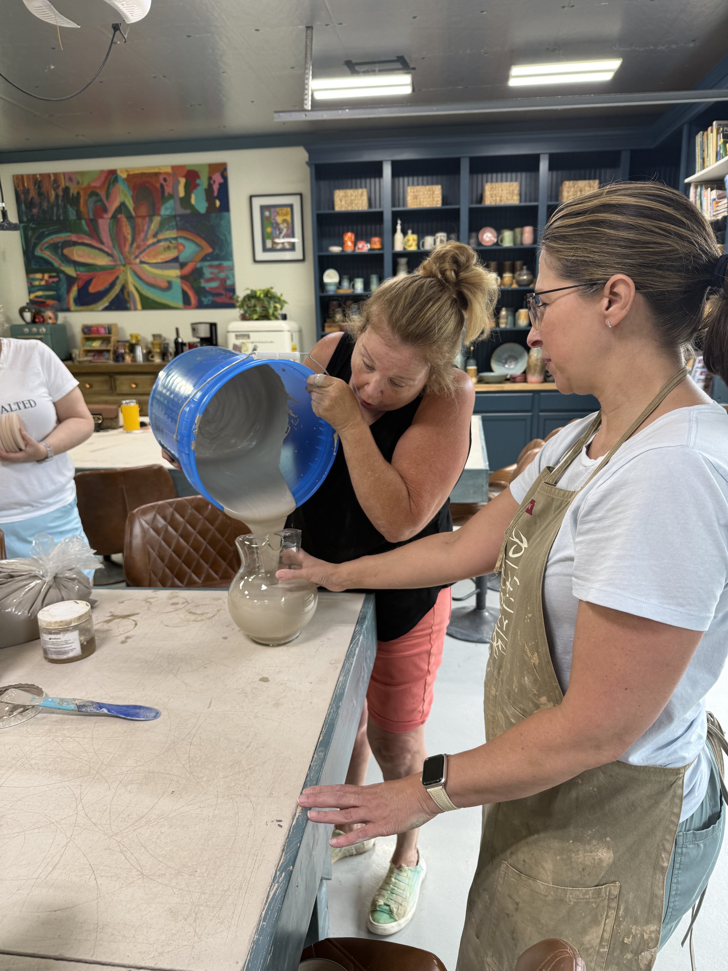 Two women pouring ceramic glaze into a pitcher in a pottery studio, with shelves of pottery in the background.