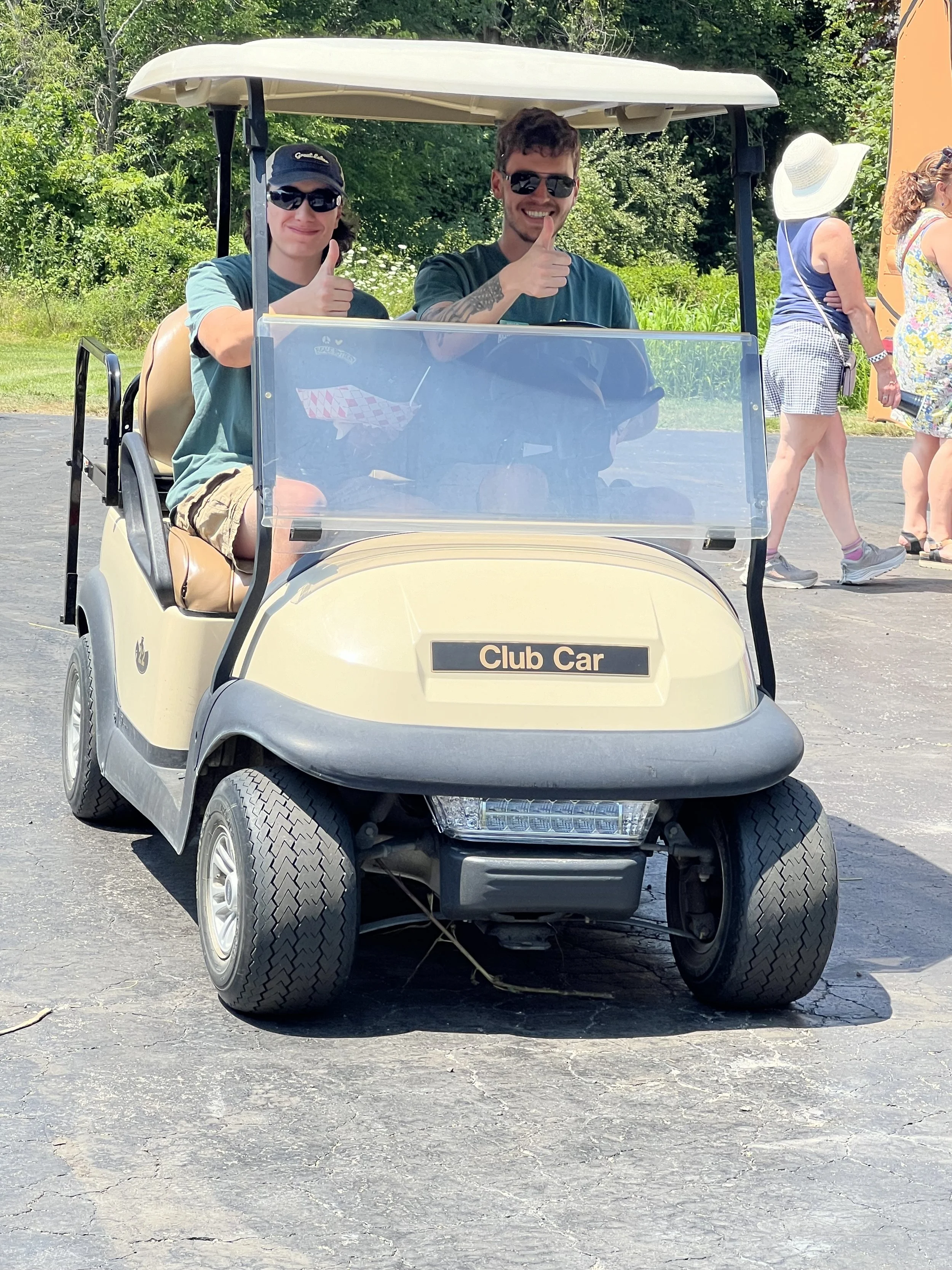 Two young men sitting in a beige golf cart labeled 'Club Car', giving a thumbs-up and smiling at the camera, with a few people walking in the background on a sunny day.