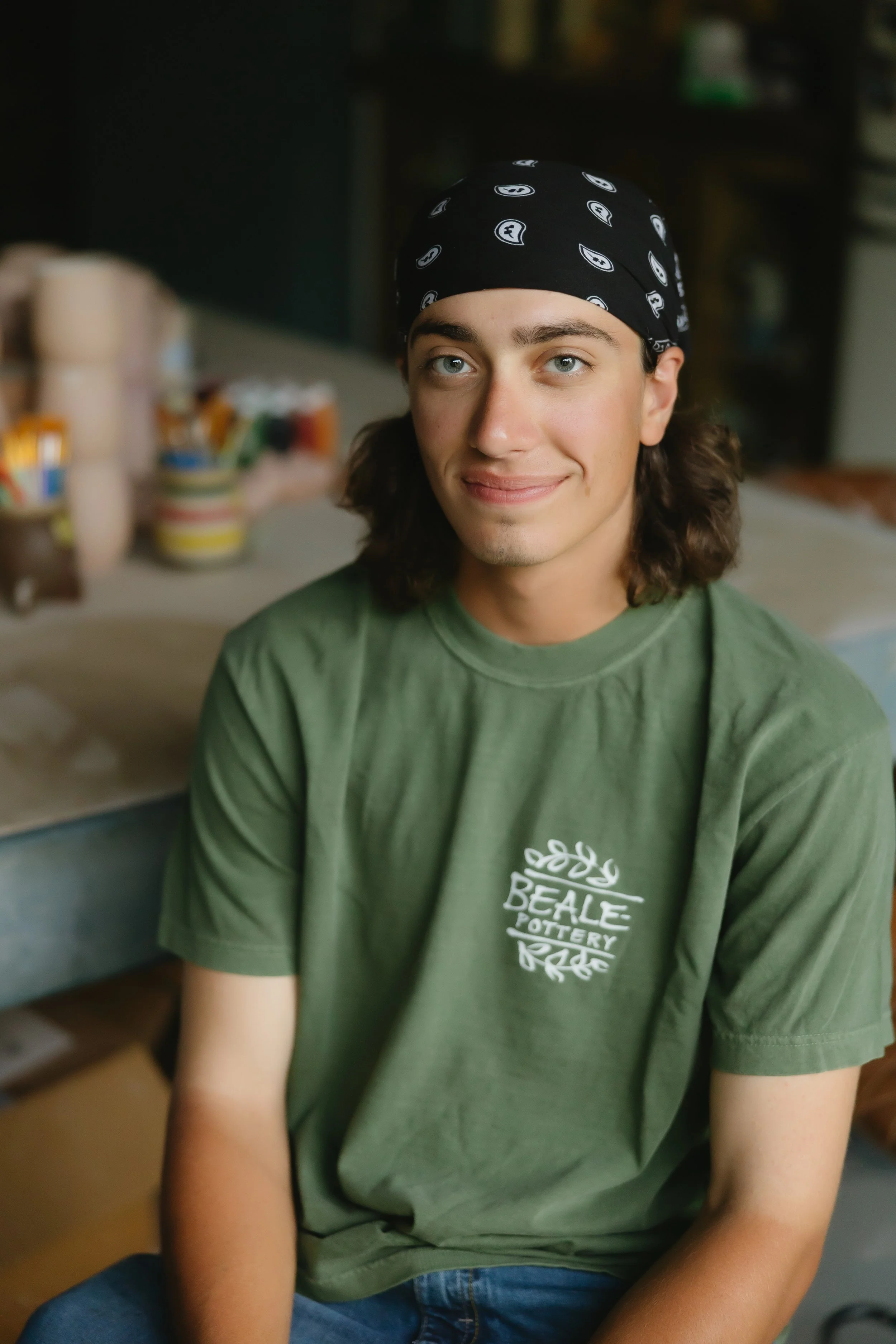 A young man with shoulder-length brown hair, wearing a black bandana and a green t-shirt with 'Beale Lottery' written on it, sitting in a casual indoor setting.