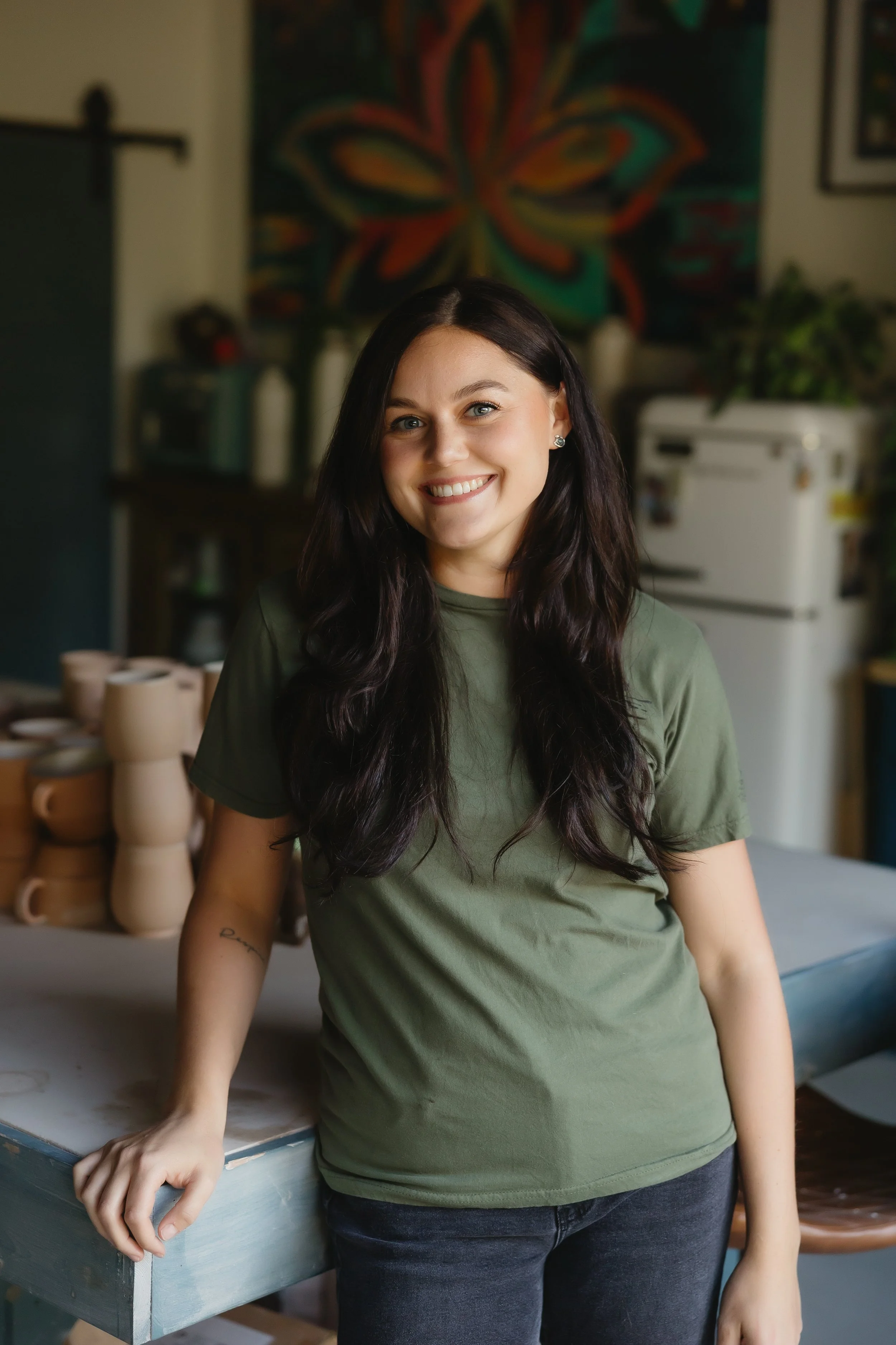 A smiling woman with long dark hair, wearing a green t-shirt and black pants, standing in a room with a colorful abstract art piece and various kitchen items in the background.