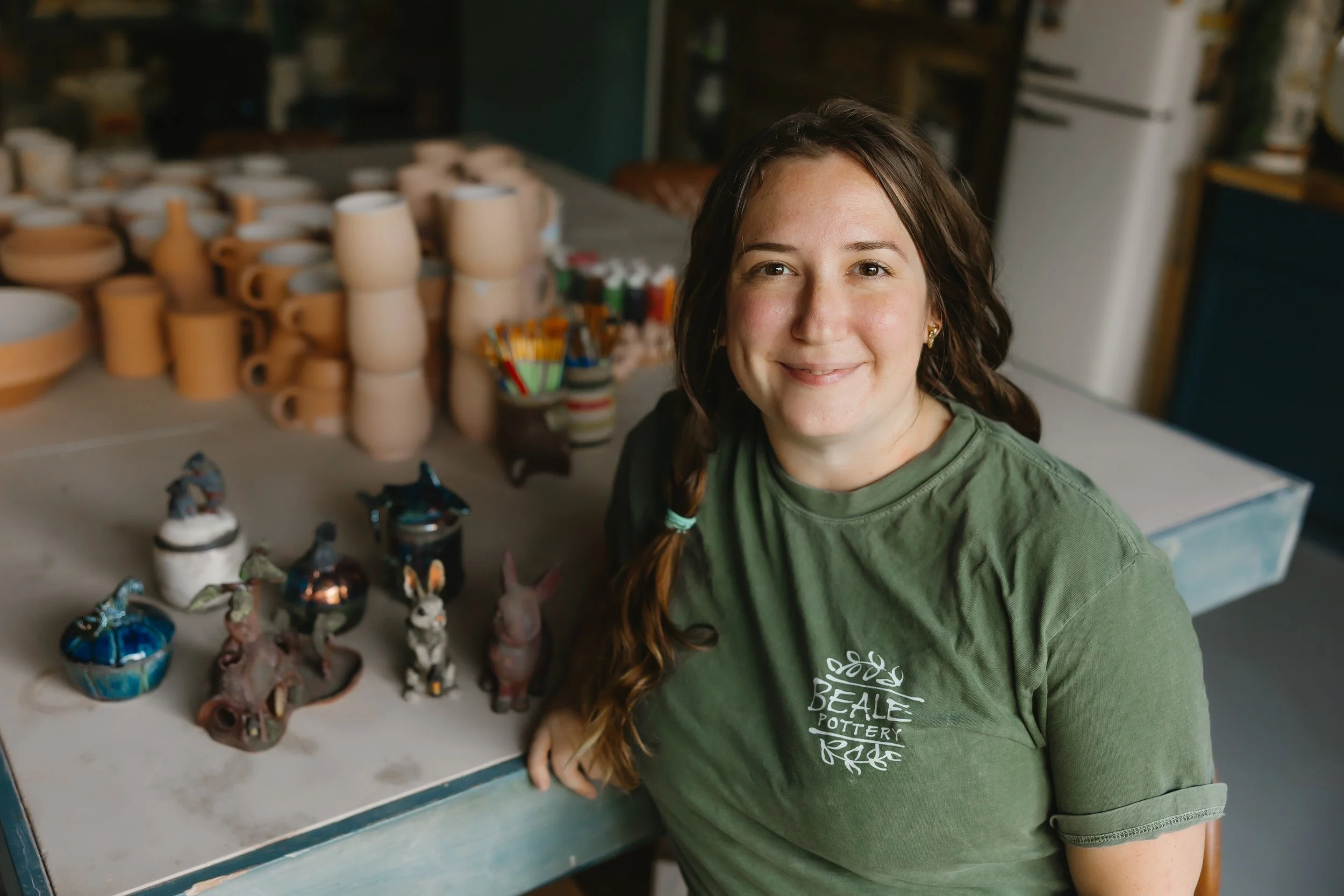 A woman with brown hair and a green Beaute Pottery t-shirt sitting at a pottery workspace with pottery pieces and art supplies on the table.