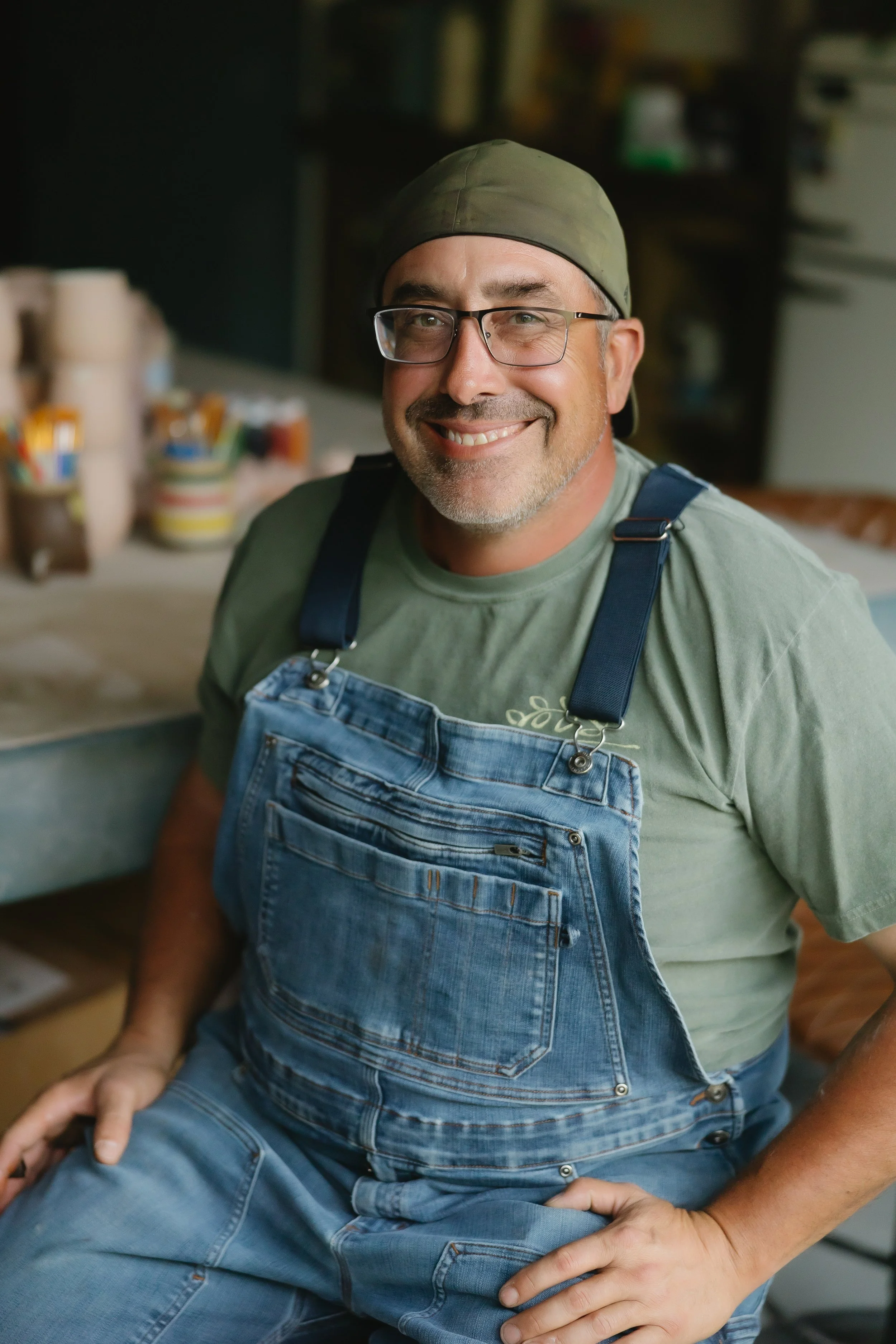 Smiling man wearing glasses, a green cap, a t-shirt, and denim overalls sitting in a workshop with art supplies behind him.