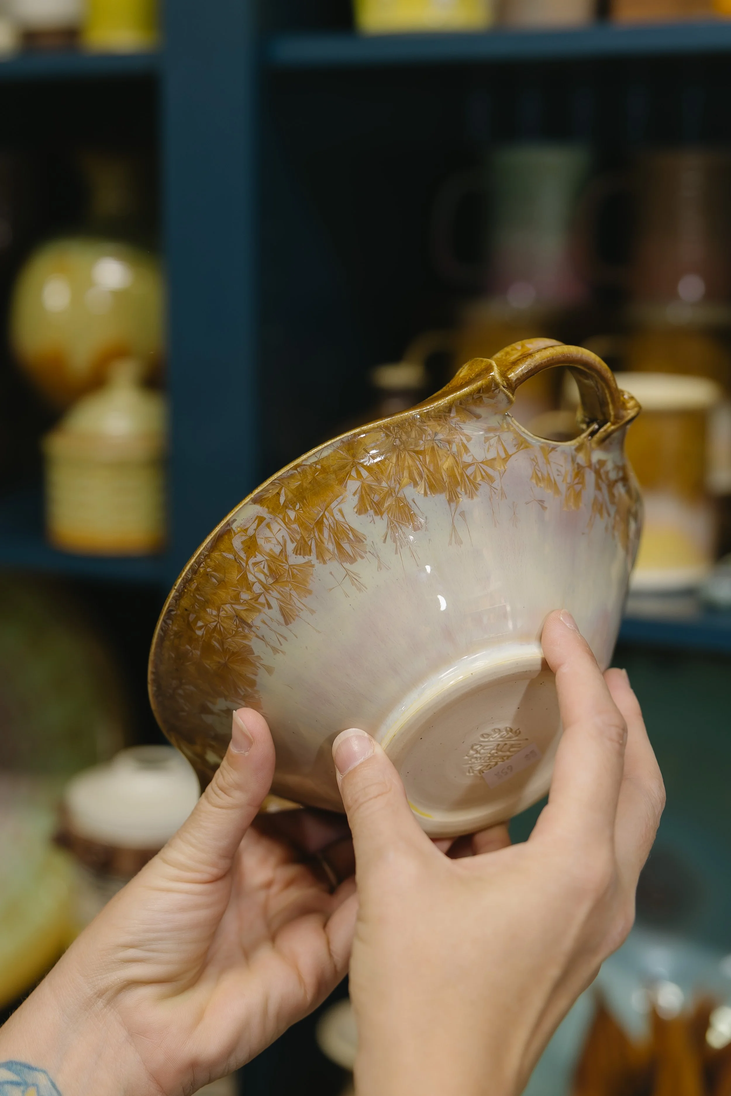 Person holding a decorative ceramic bowl with a brown and cream glazed finish, featuring a tree and leaf pattern around the rim, in a store with other pottery items on shelves in the background.