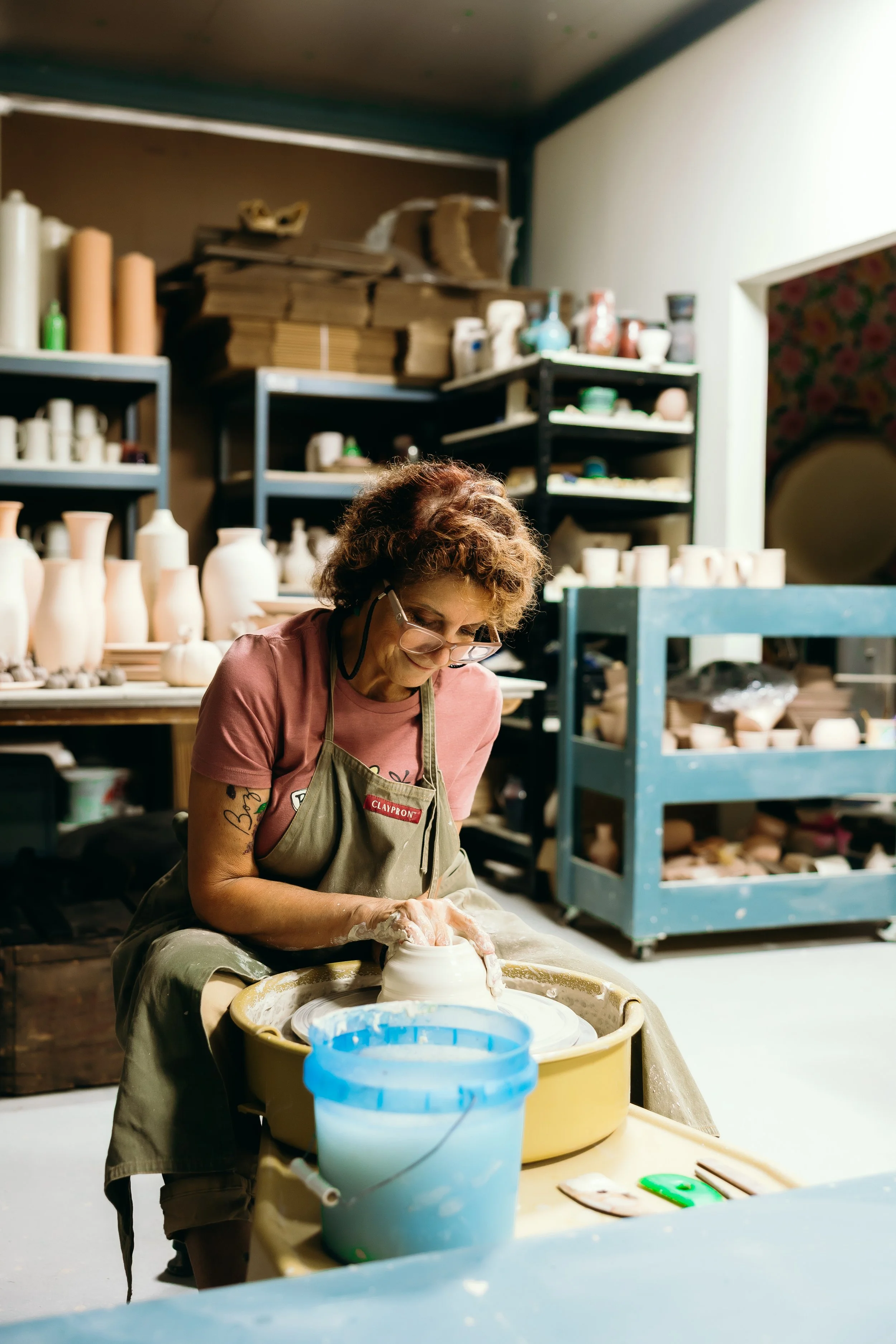 A woman working on pottery in a ceramics studio. She is shaping a clay piece on a pottery wheel with a bucket of water nearby. Shelves behind her hold finished and in-progress ceramic pieces.