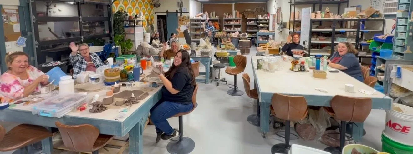 A group of people sitting around tables in a ceramics studio, smiling and waving at the camera. The tables are filled with clay, tools, and ceramics projects. Shelves in the background hold various pottery supplies and finished pieces.