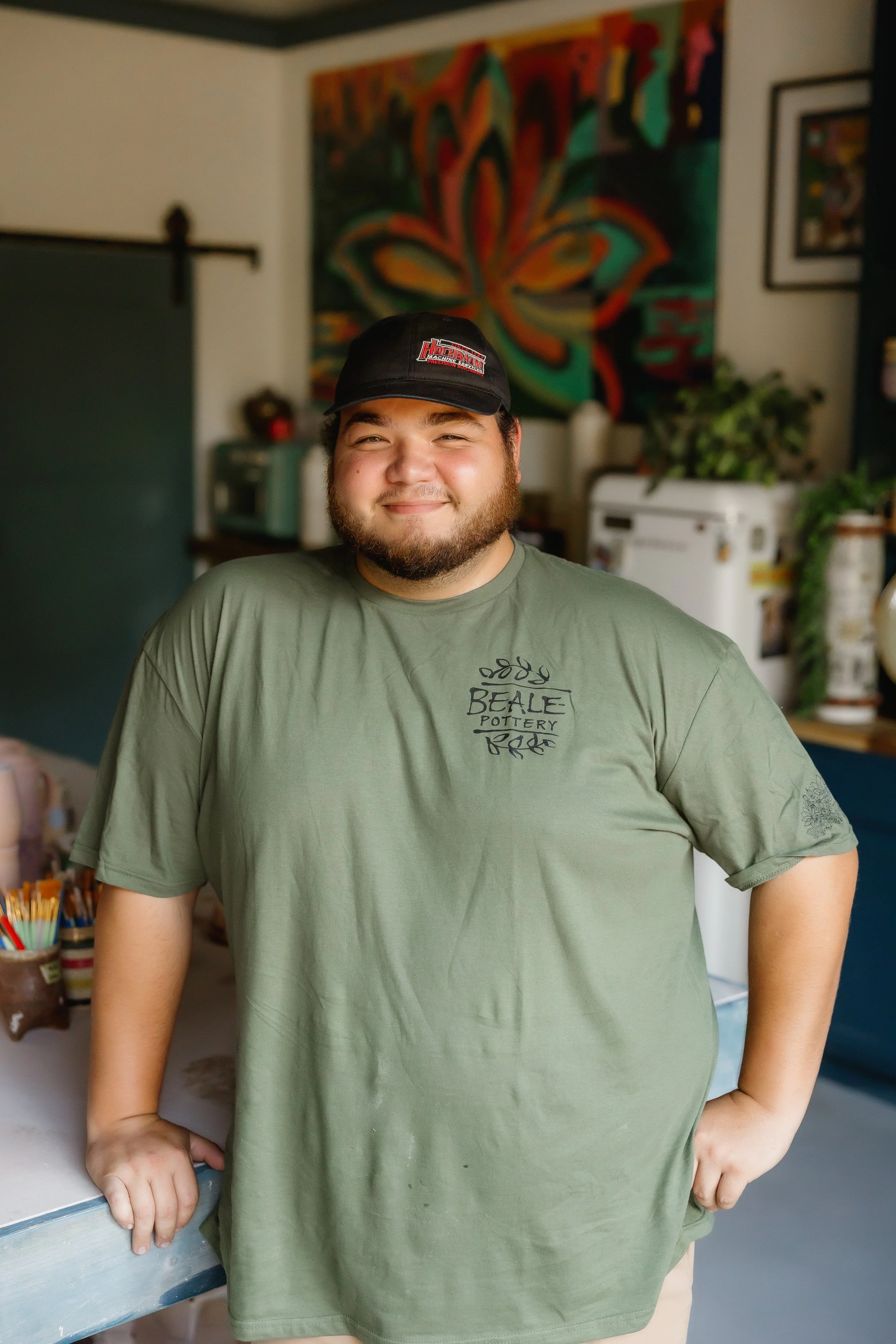 A young man with a beard and a baseball cap, wearing a green T-shirt, smiling and leaning on a table inside a room decorated with colorful artwork and plants.