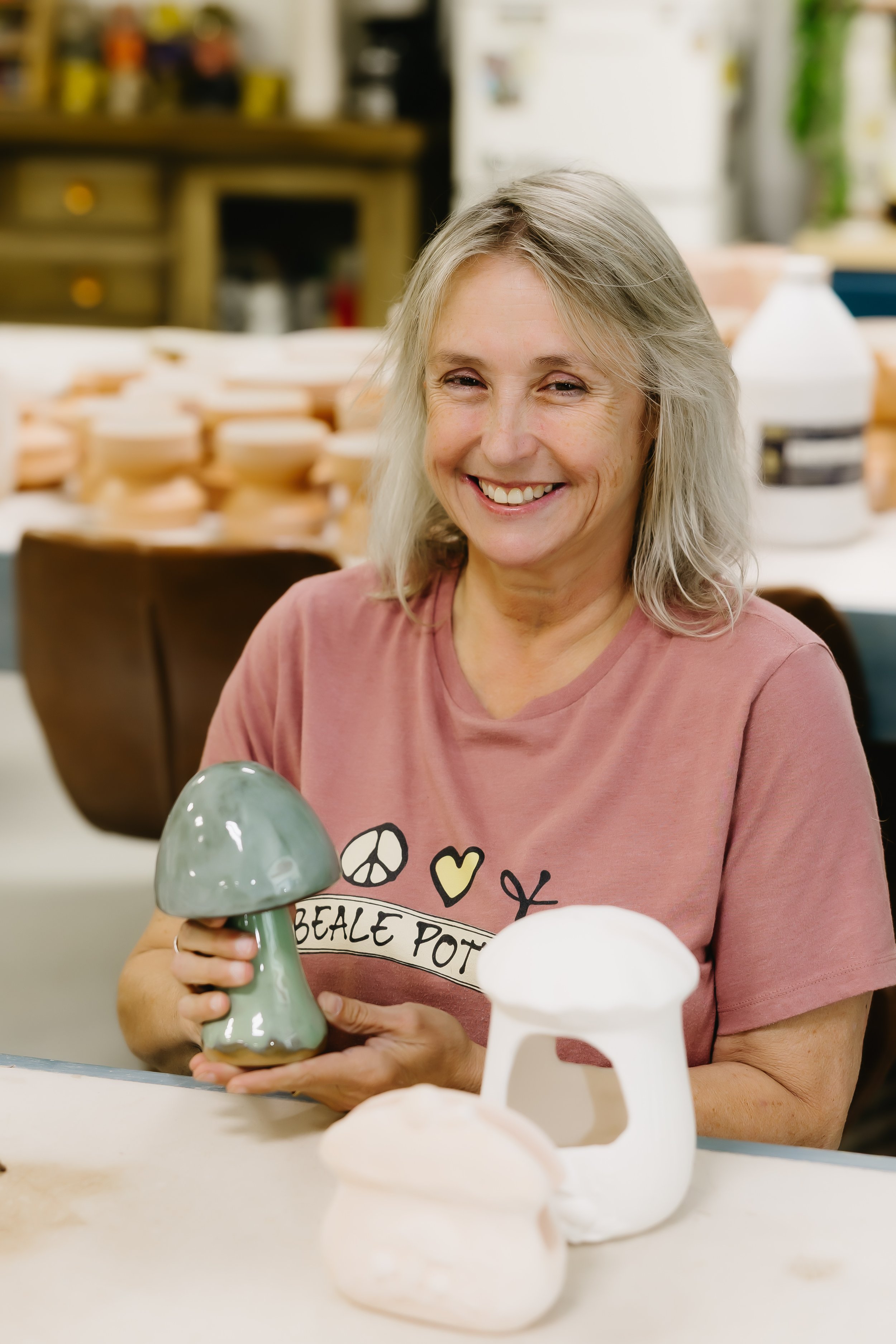 A woman smiling and holding a ceramic mushroom sculpture, with other ceramic items on the table in front of her, in a ceramics workshop or studio.