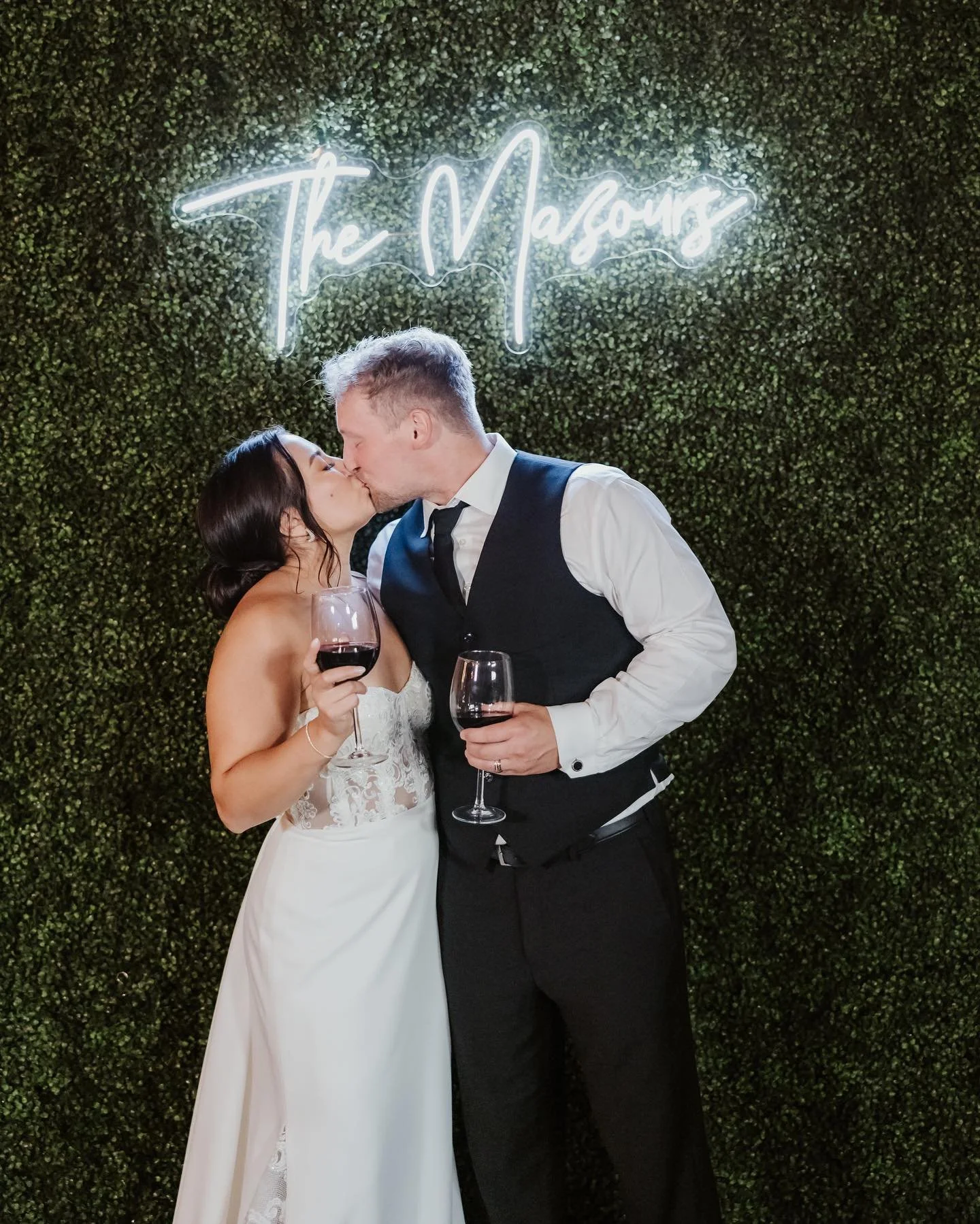 bride and groom kissing in front of boxwood wall with neon sign of their last name