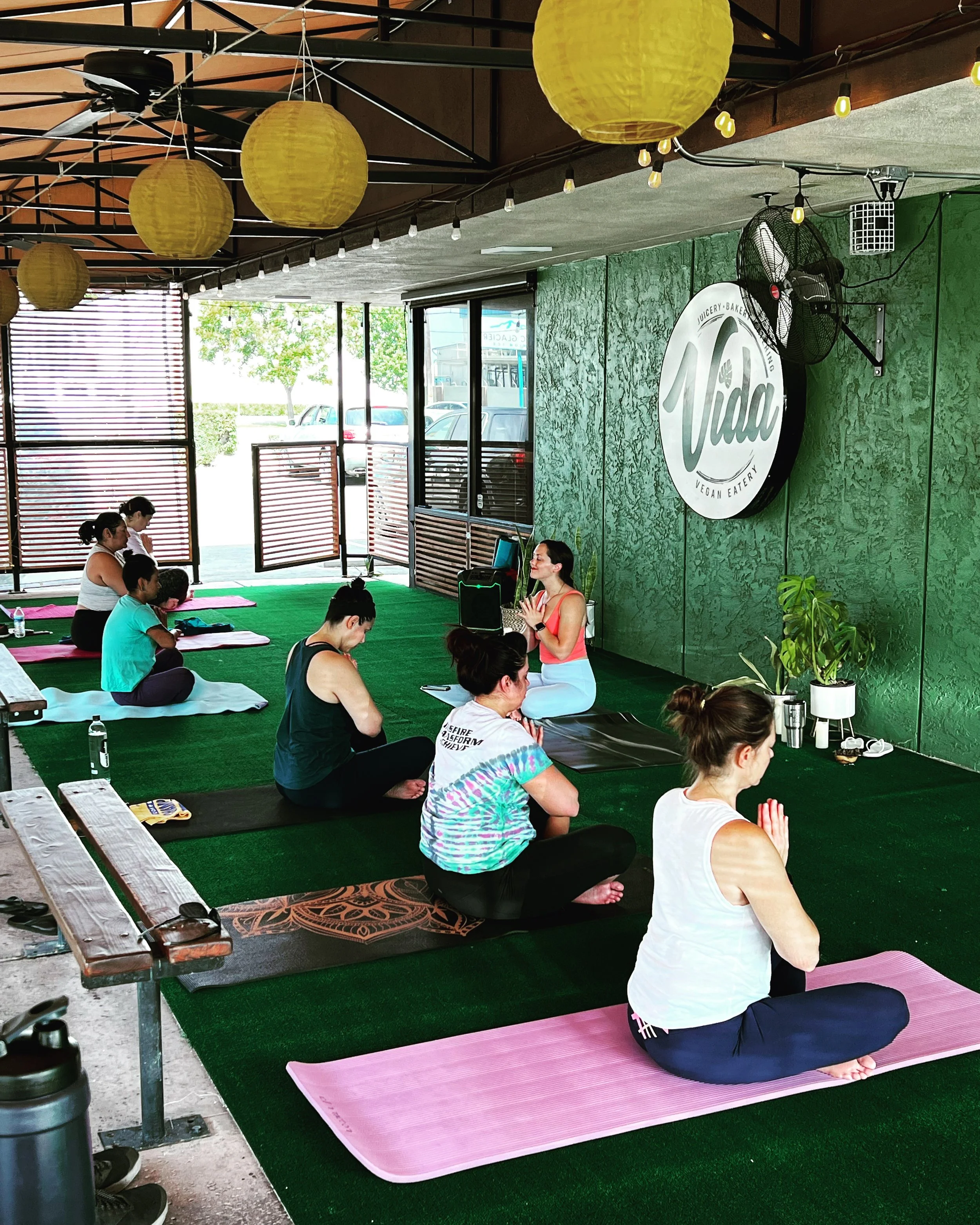 Group yoga class with participants seated on mats in an outdoor space, green carpet, and yellow lanterns, near a sign reading 'Vida.'