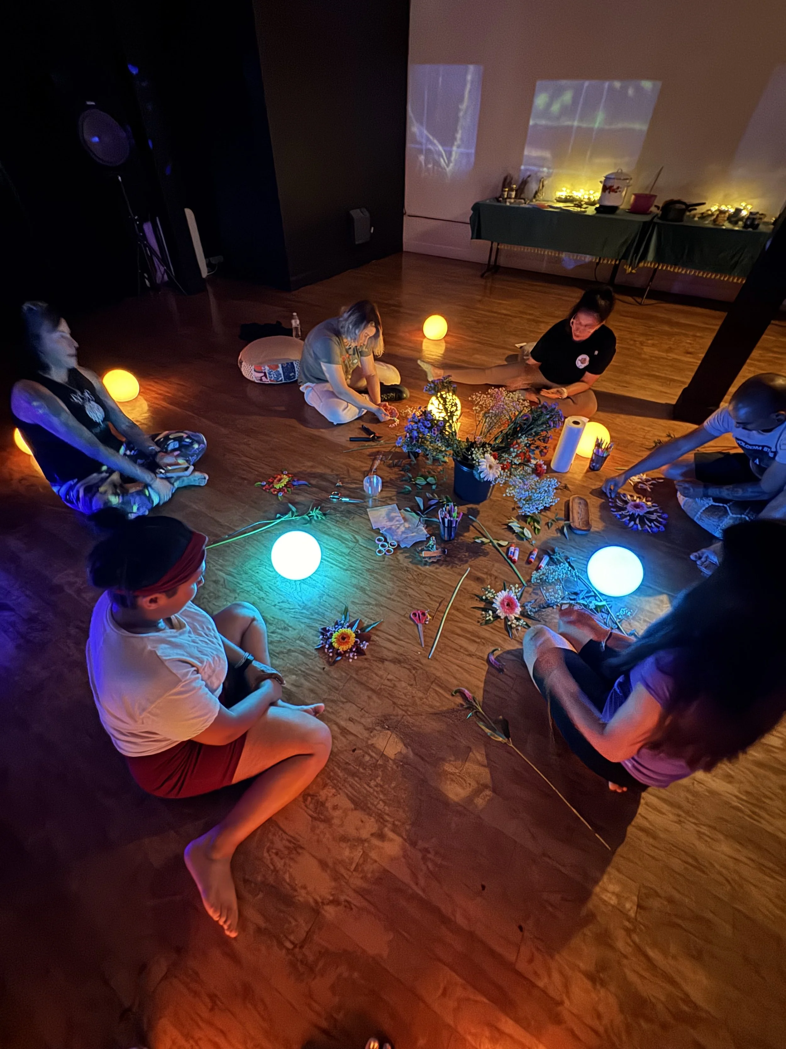 People seated on the floor, creating floral arrangements with various flowers and tools, illuminated by colored lights in a dim room.