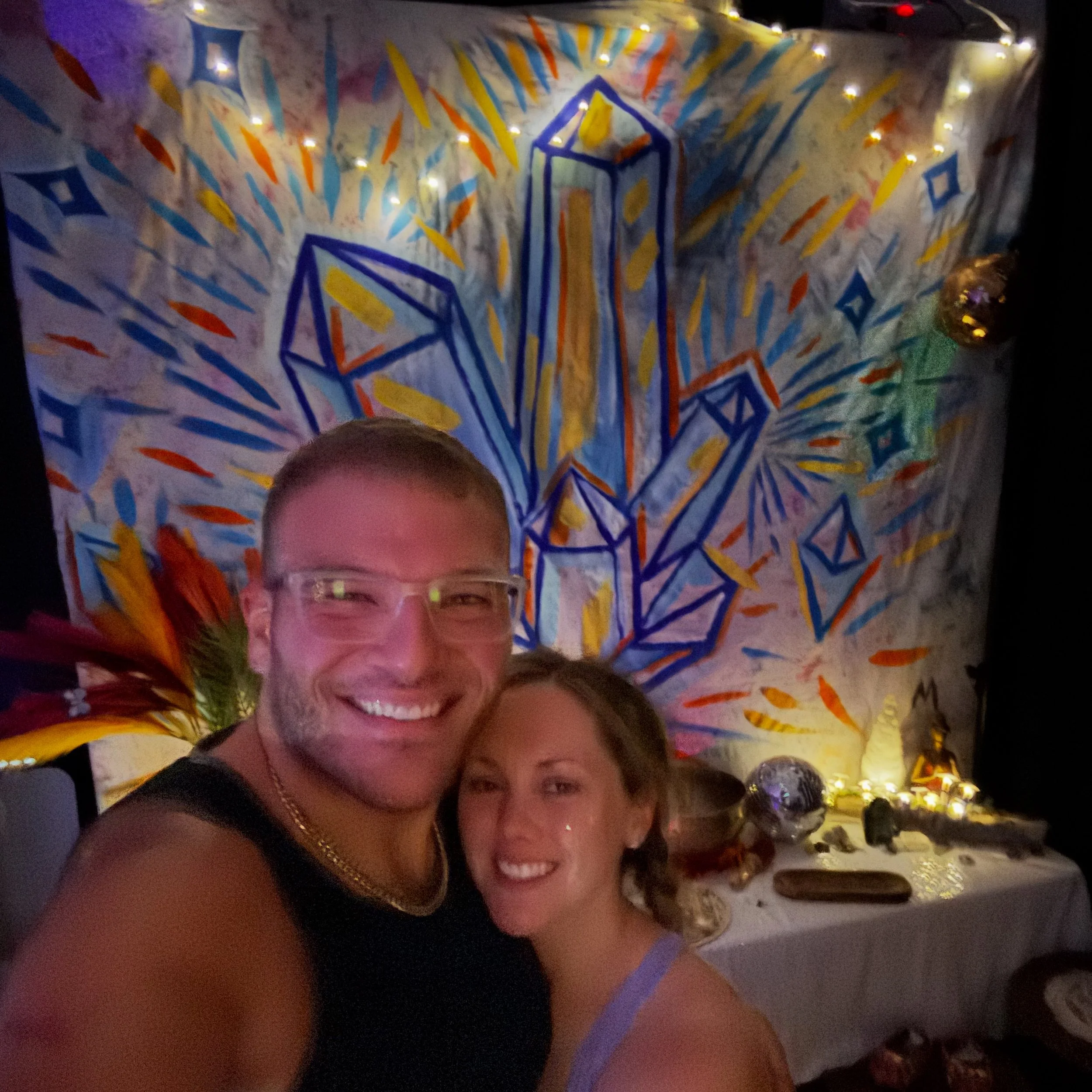 A smiling man and woman taking a selfie together at a celebration, with a colorful abstract mural and a table with decorations and lights in the background.