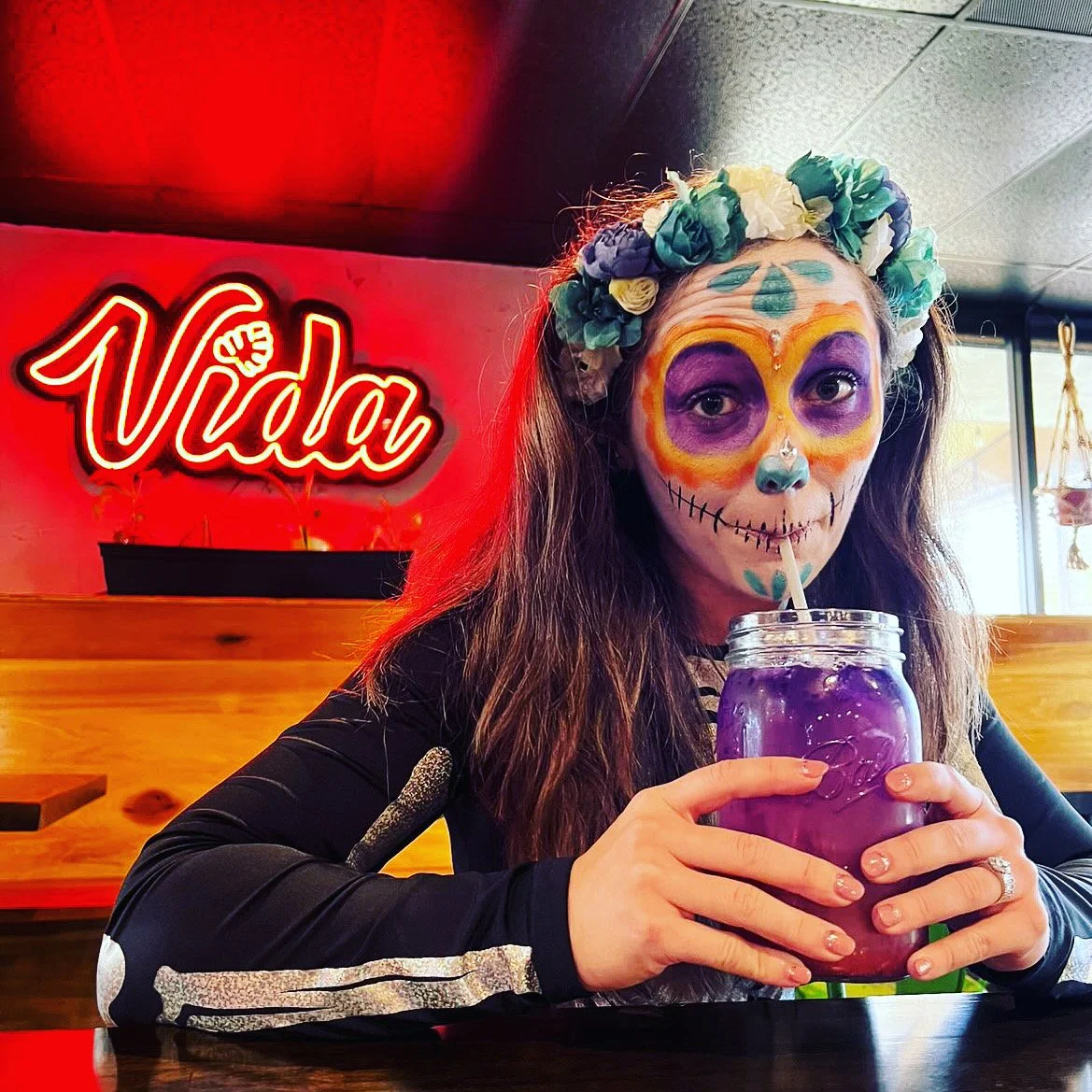 Person with Day of the Dead face paint and flower crown drinking from a mason jar in front of a neon "Vida" sign.