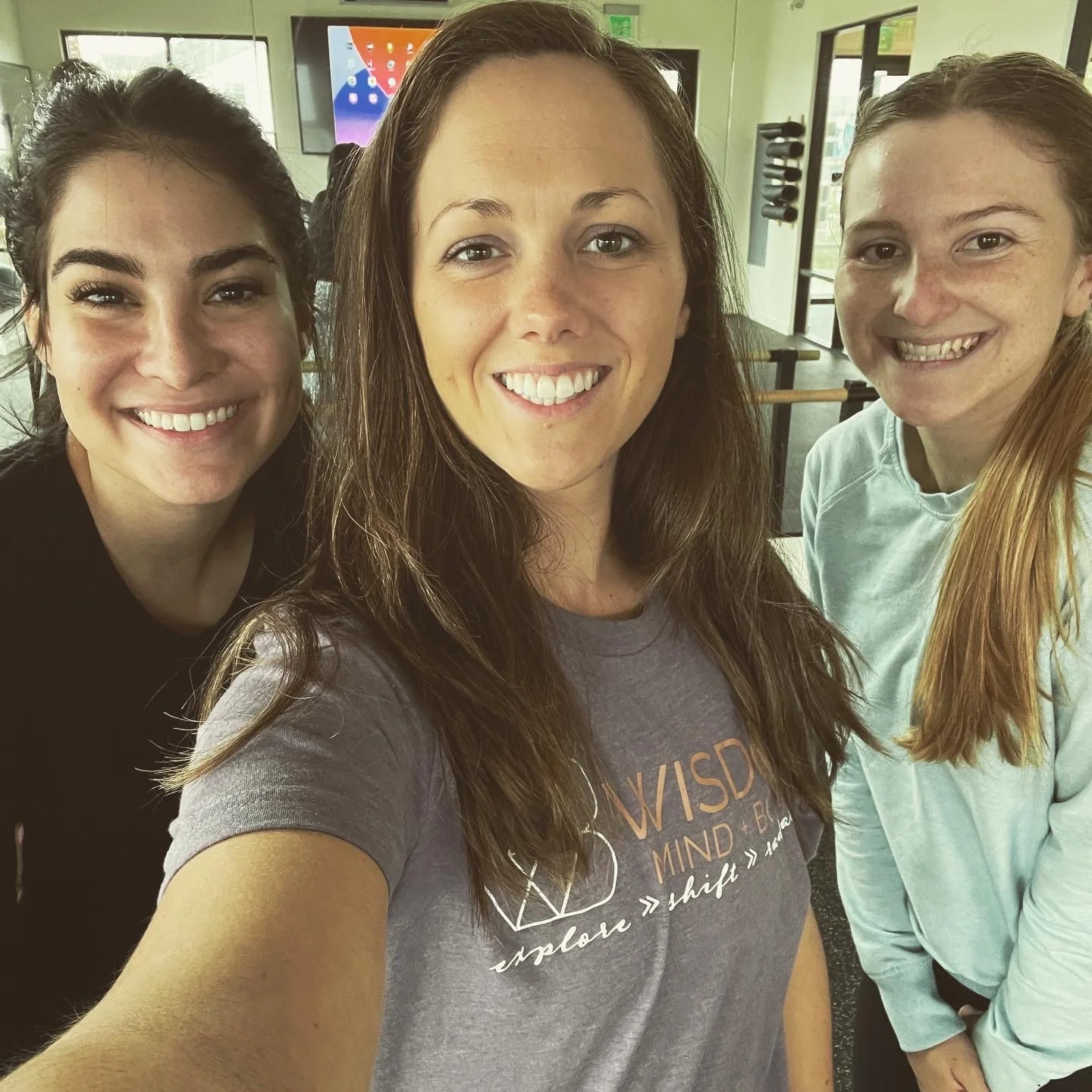 Three women smiling indoors, one wearing a shirt with "Wisdom Mind-Body" written on it, standing in a room with exercise equipment.