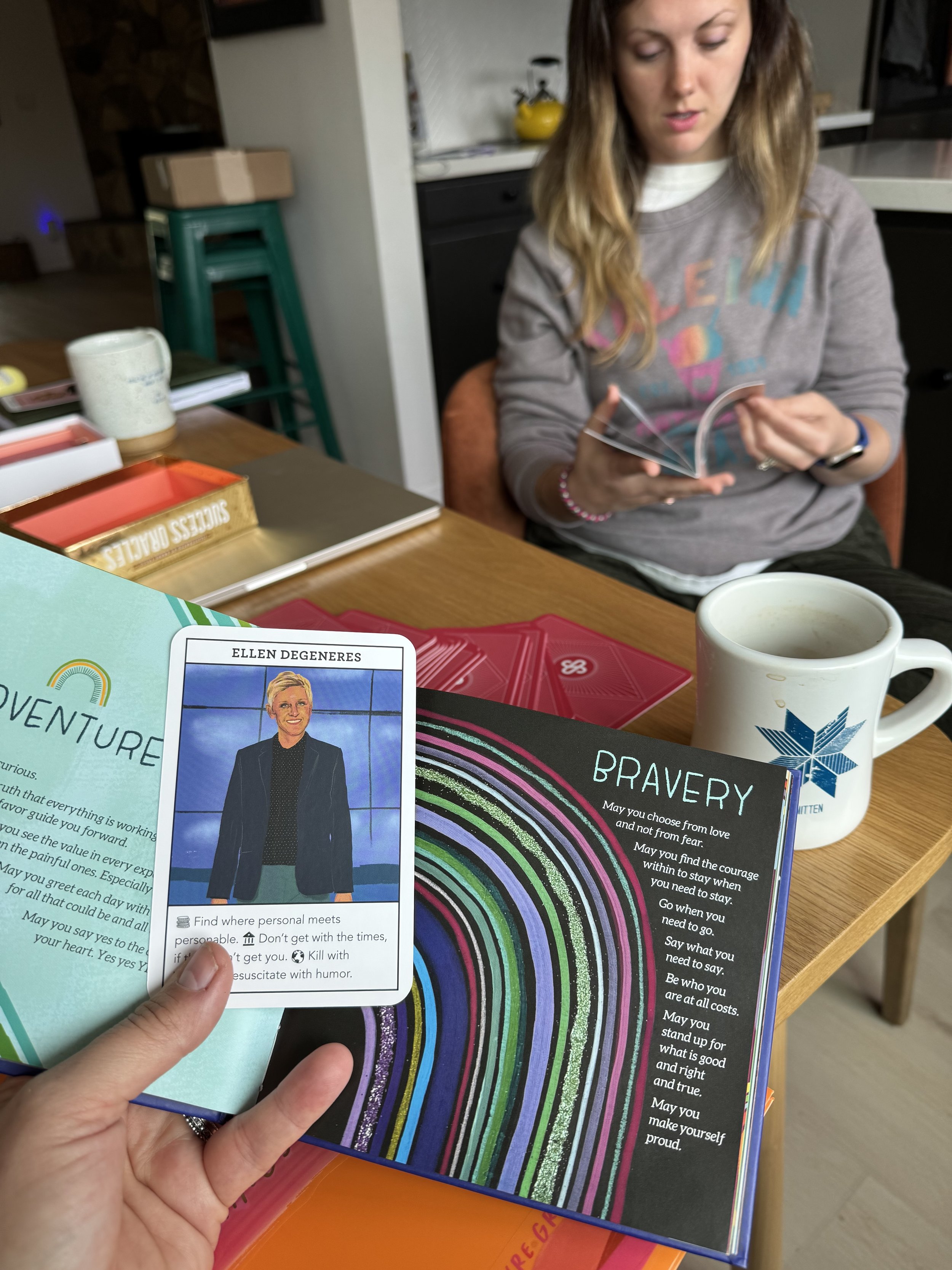 A person holding a card with Ellen DeGeneres's picture and a motivational card with the word 'Bravery' and a rainbow-like design on a wooden table with a woman reading a booklet and a coffee mug nearby.